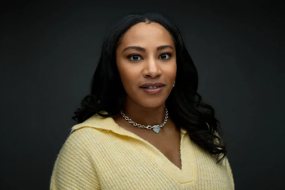 A woman with long dark hair wearing a yellow sweater and silver necklace with a heart pendant, posing against a dark background.