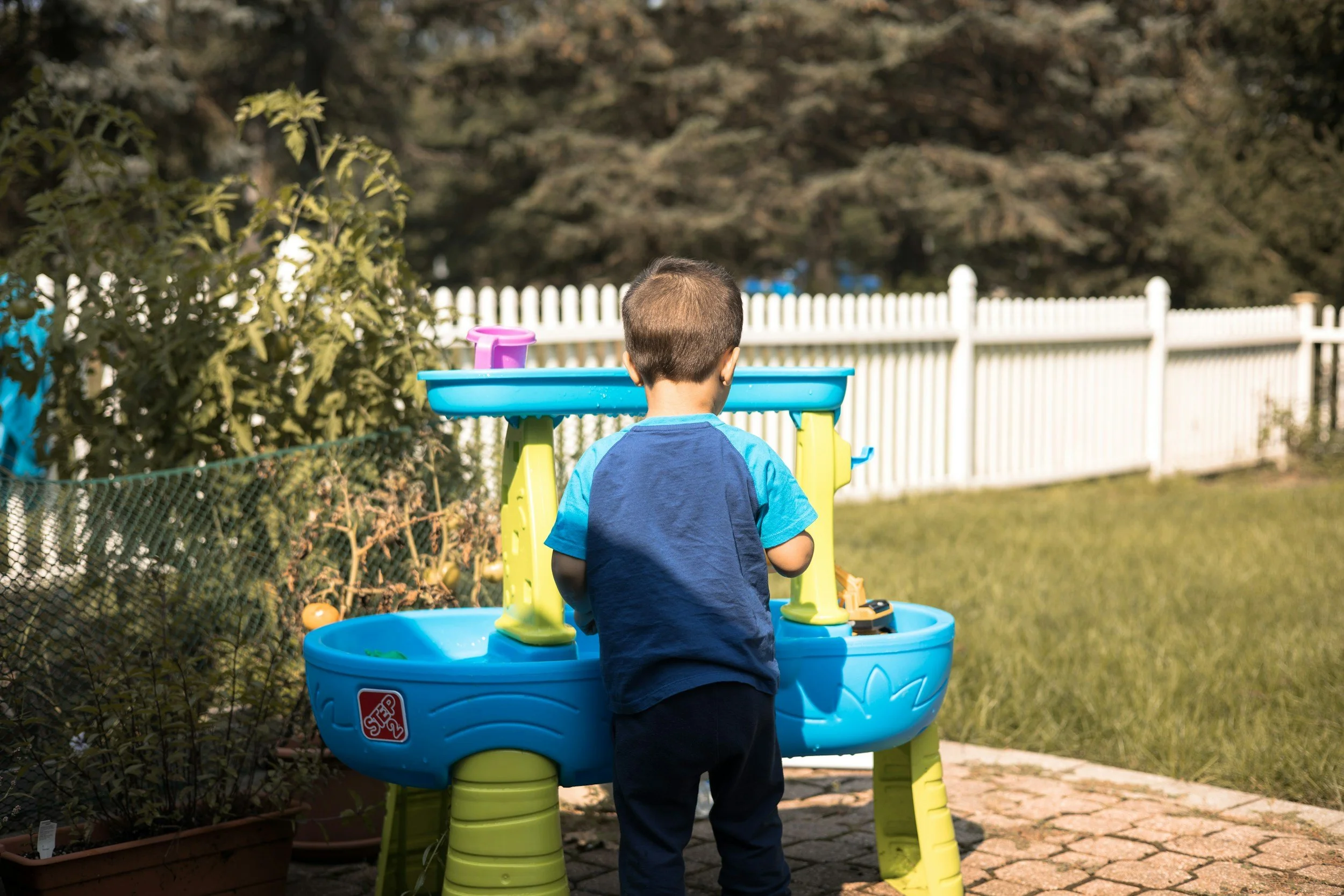 A young boy with short hair wearing a blue shirt is standing in front of a blue plastic water table outdoors. The water table contains water and toy accessories. The background includes a white picket fence, trees, and plants, suggesting a backyard or garden setting.