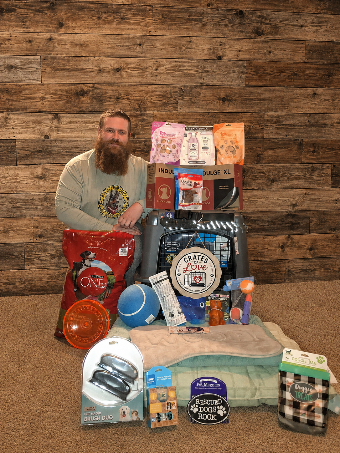 A man with a beard and long hair standing next to a pile of various pet supplies, including dog food, toys, grooming items, and a pet crate, with a wooden wall background.