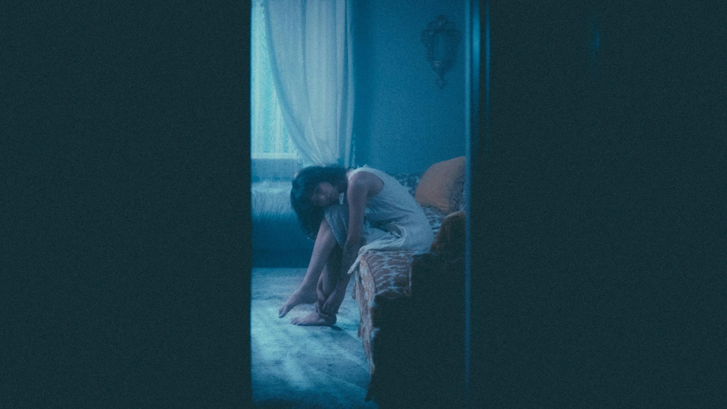 Woman sitting on a bed, bending forward, in a dimly lit room with blue curtains, visible through a doorway.