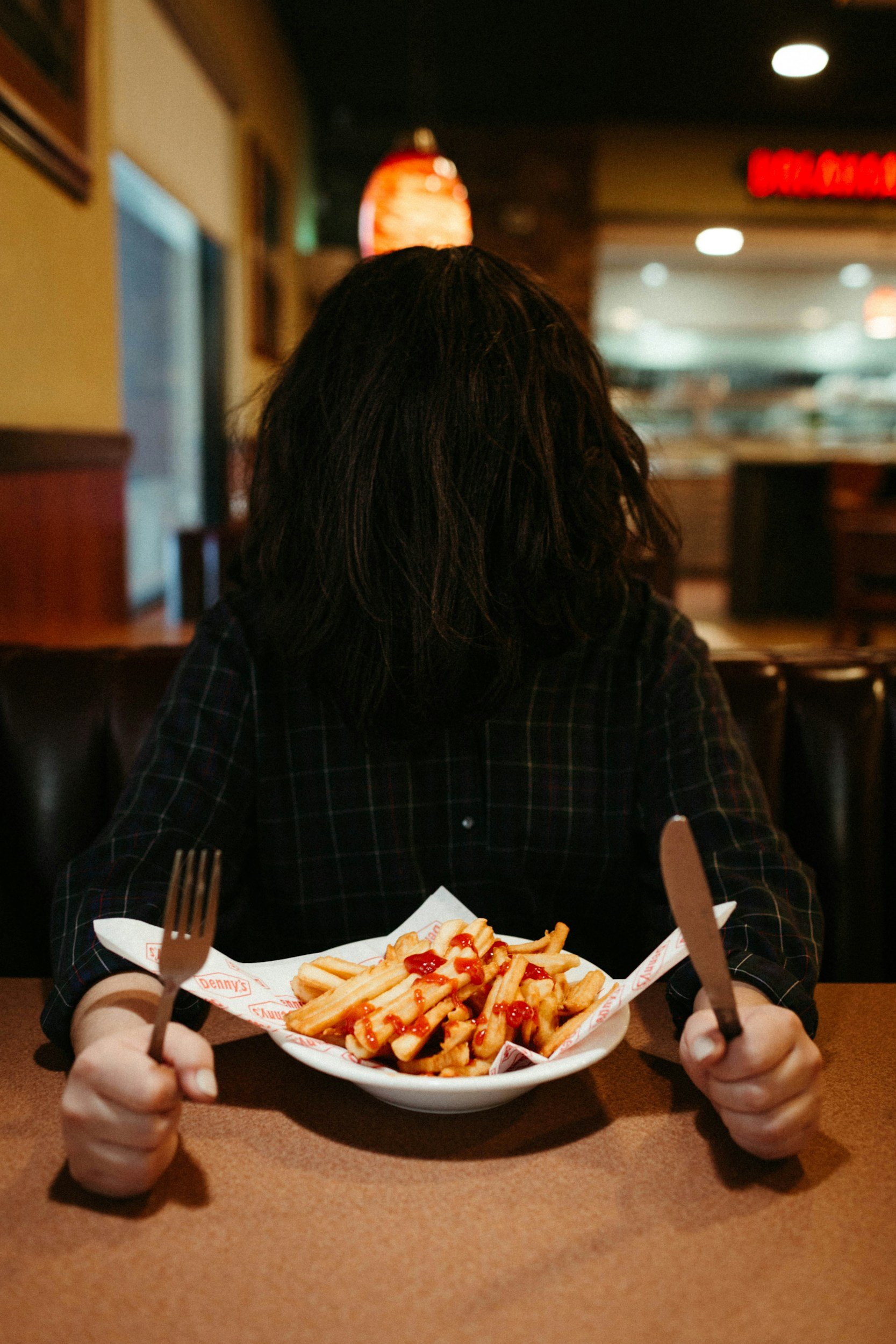 A person with long dark hair sitting at a restaurant table, holding a plate of fries topped with ketchup, with a fork in one hand and a knife in the other.