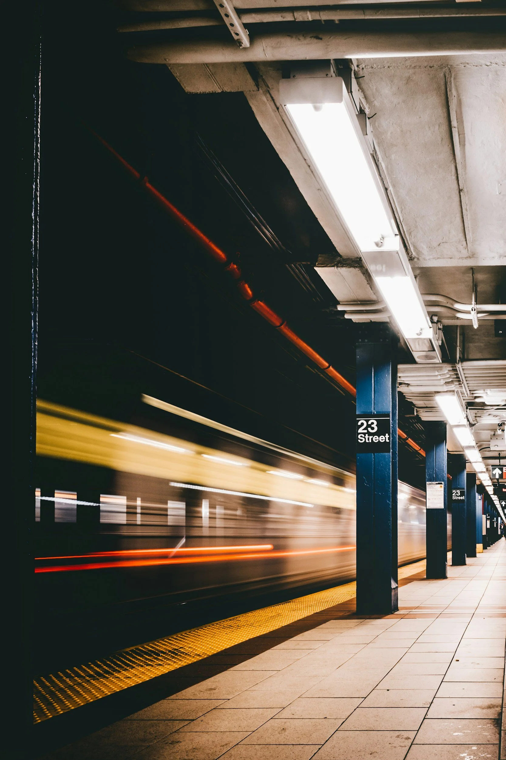 A subway station platform with a train passing through at high speed, creating blurred streaks of yellow and red lights. The platform has black support columns with signs that read "23 Street." The ceiling has fluorescent lighting and exposed pipes.