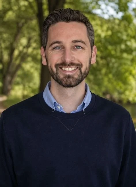 A smiling man with a beard and brown hair outdoors with green trees in the background, wearing a dark sweater over a collared shirt.