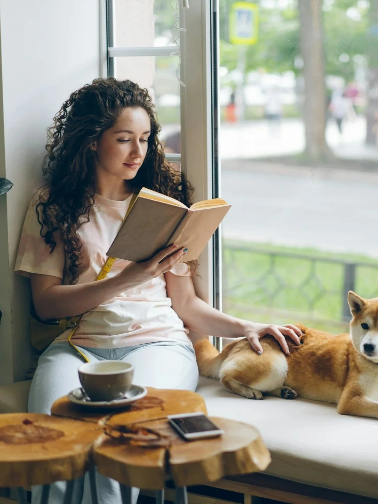 A woman with curly hair reading a book while sitting by a window, petting a Shiba Inu dog, with a cup and saucer on a wooden table nearby.