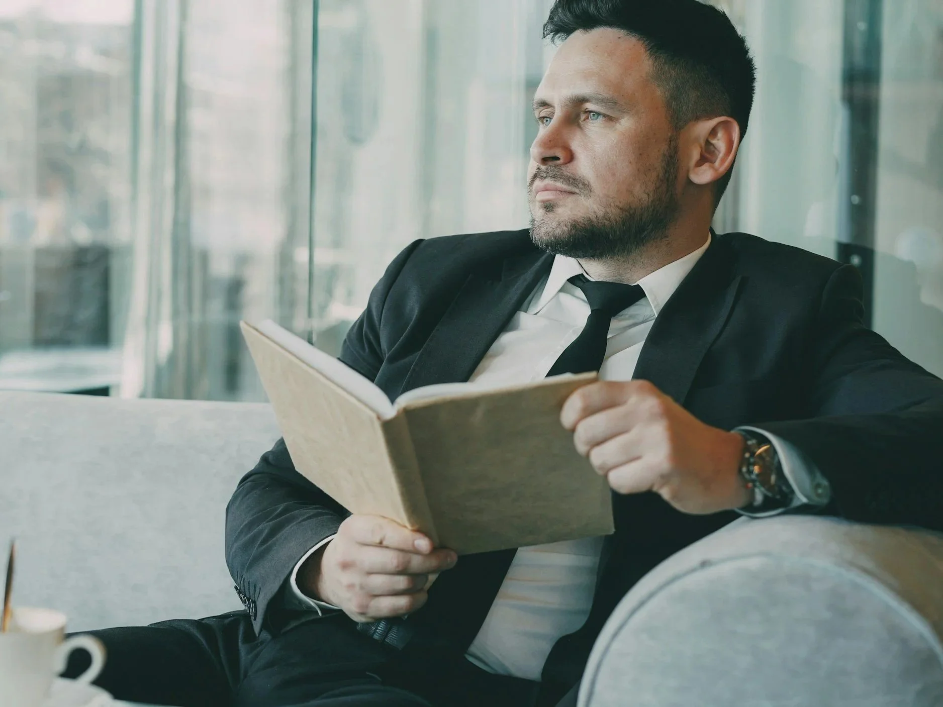 A man in a business suit, with dark hair and a beard, is sitting on a gray sofa, holding an open newspaper, and looking thoughtfully to his right. He is in a modern office or public space with glass windows in the background.