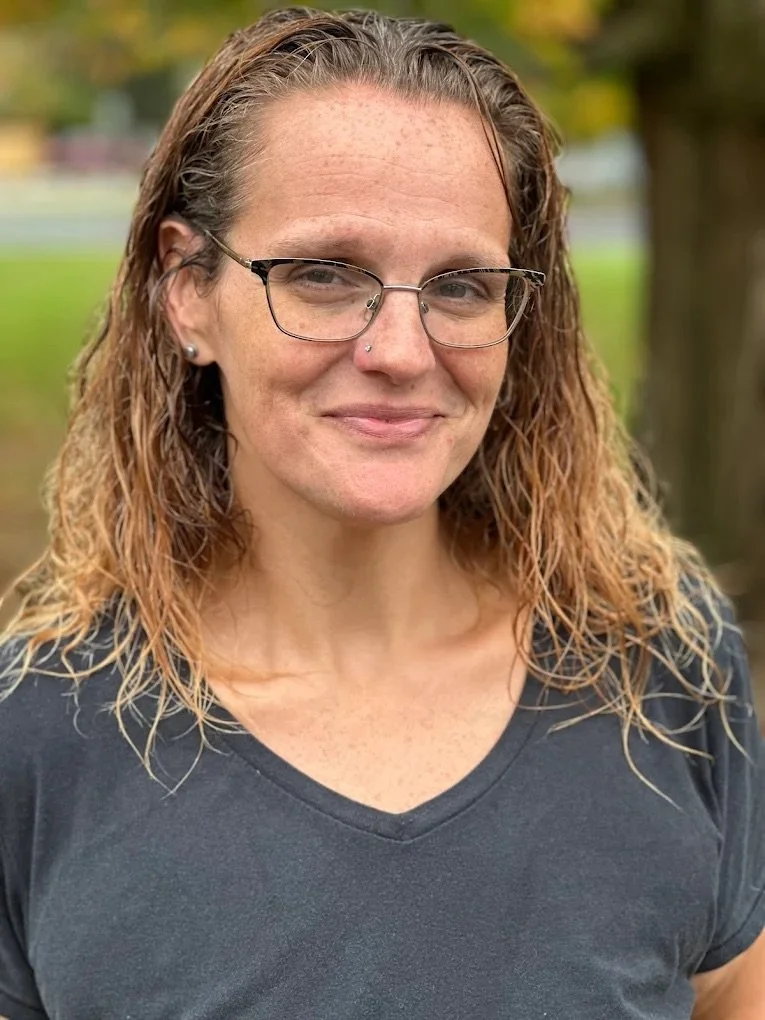 A woman with curly, shoulder-length hair, glasses, and a nose piercing, standing outdoors in front of trees and a blurred natural background, wearing a black V-neck shirt.