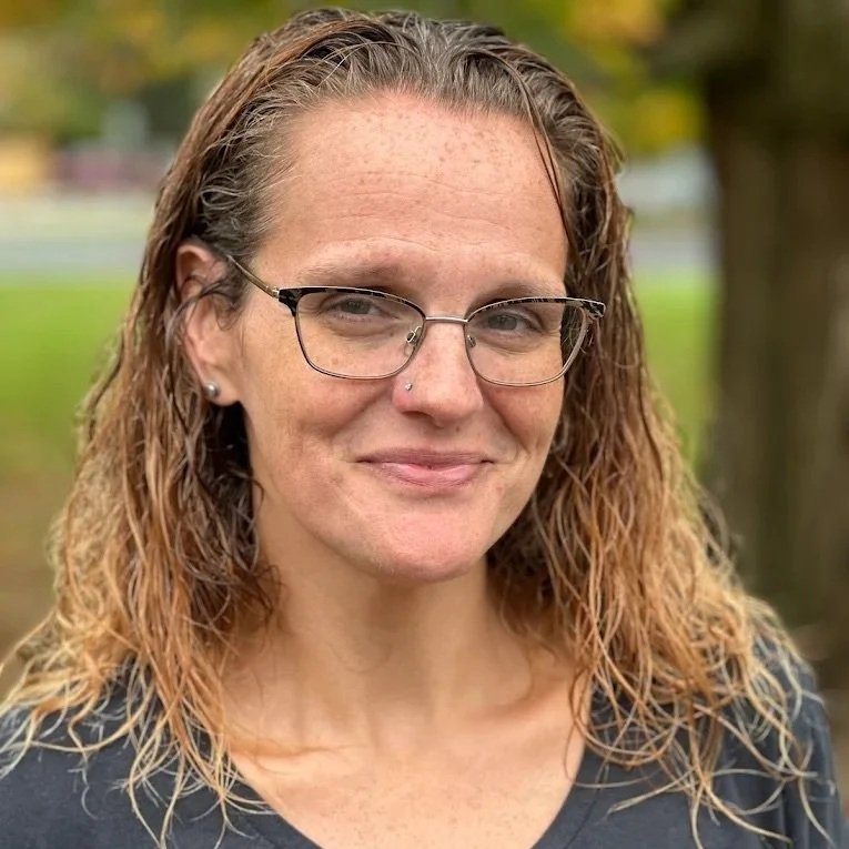 Close-up of a woman with light skin, curly light brown hair, glasses, and a nose piercing outdoors.