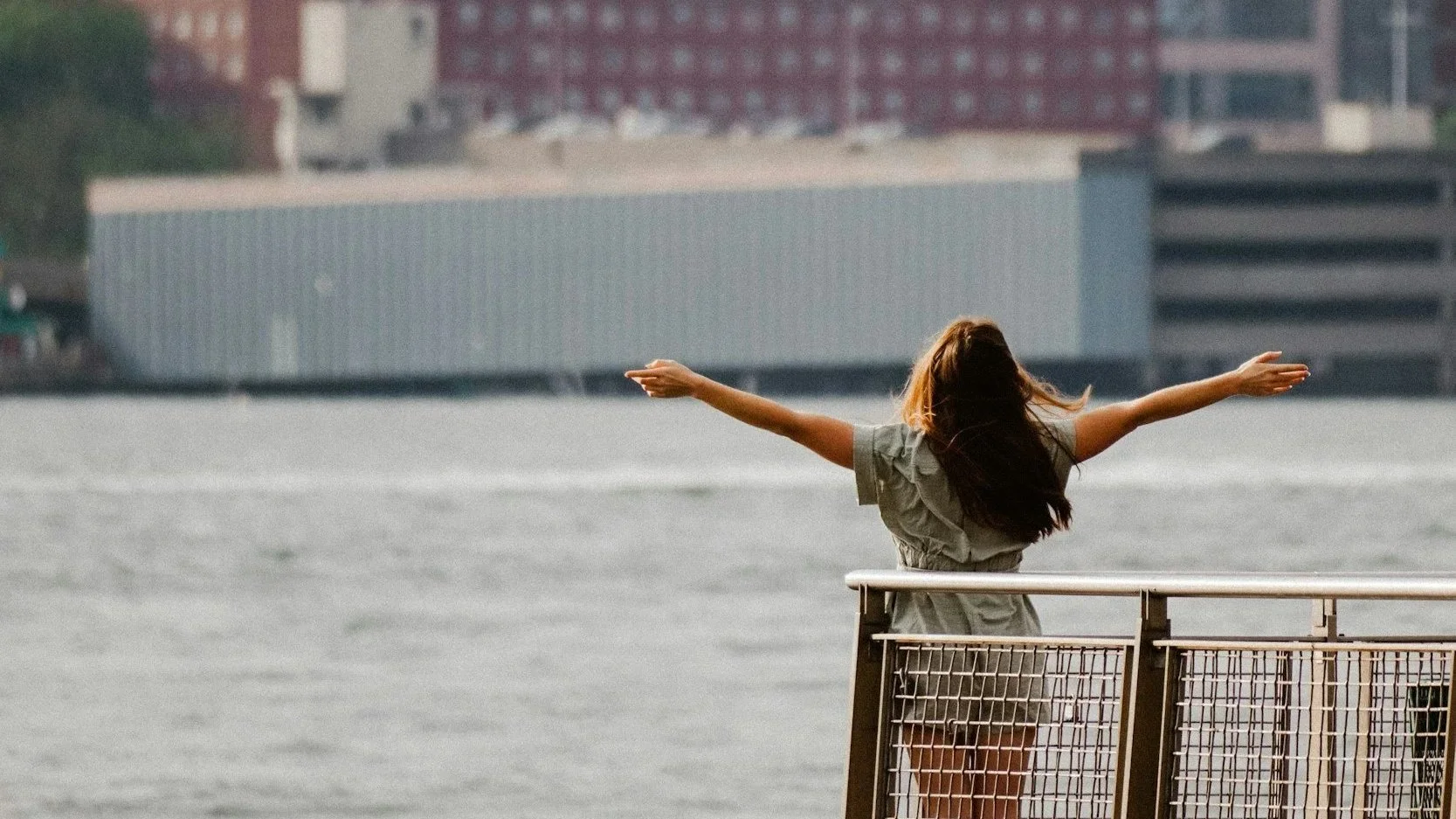 A woman with long hair in a gray dress stands on a dock or platform with her arms stretched out wide, facing a body of water, with buildings in the background.