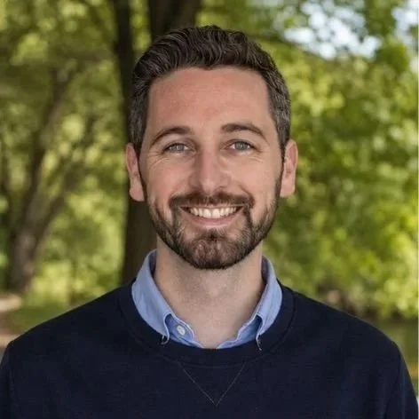 Portrait of a man with short dark hair and a beard, smiling while standing outdoors with trees and greenery in the background.