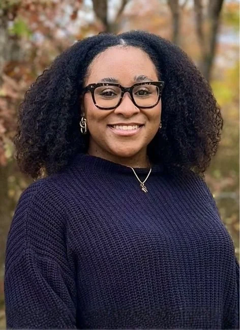 Smiling woman with curly black hair, glasses, and earrings, standing outdoors with autumn-colored trees in the background.