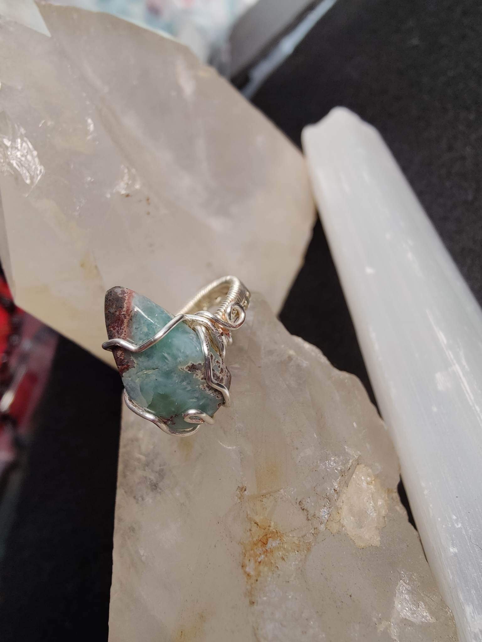 Close-up of a small turquoise gemstone wrapped in silver wire, resting on a rough surface, with a black object and white pipe in the background.