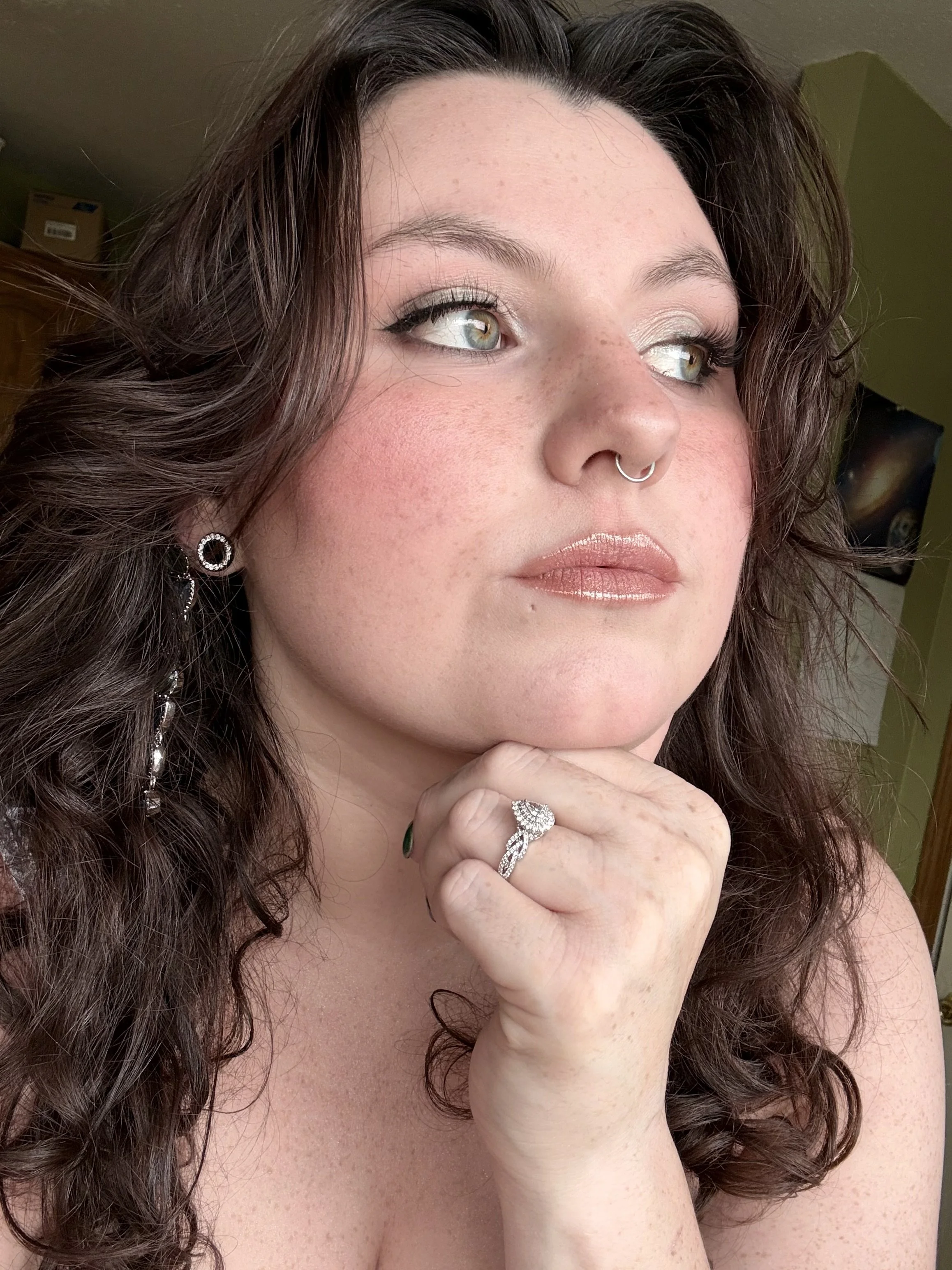 A close-up of a woman with curly brown hair, wearing a nose ring, earrings, and a ring on her finger. She has light makeup and is resting her chin on her hand.