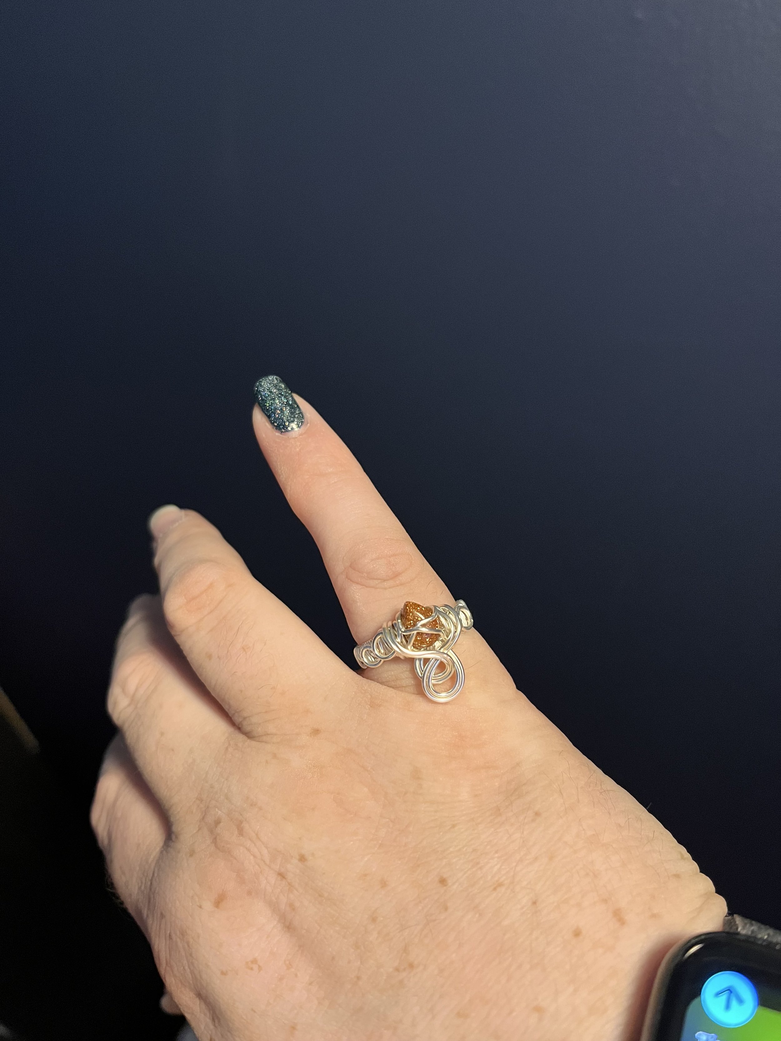 A person's hand wearing a silver ring with a gold and silver design, a black watch, and painted fingernails with glittery dark green nail polish.