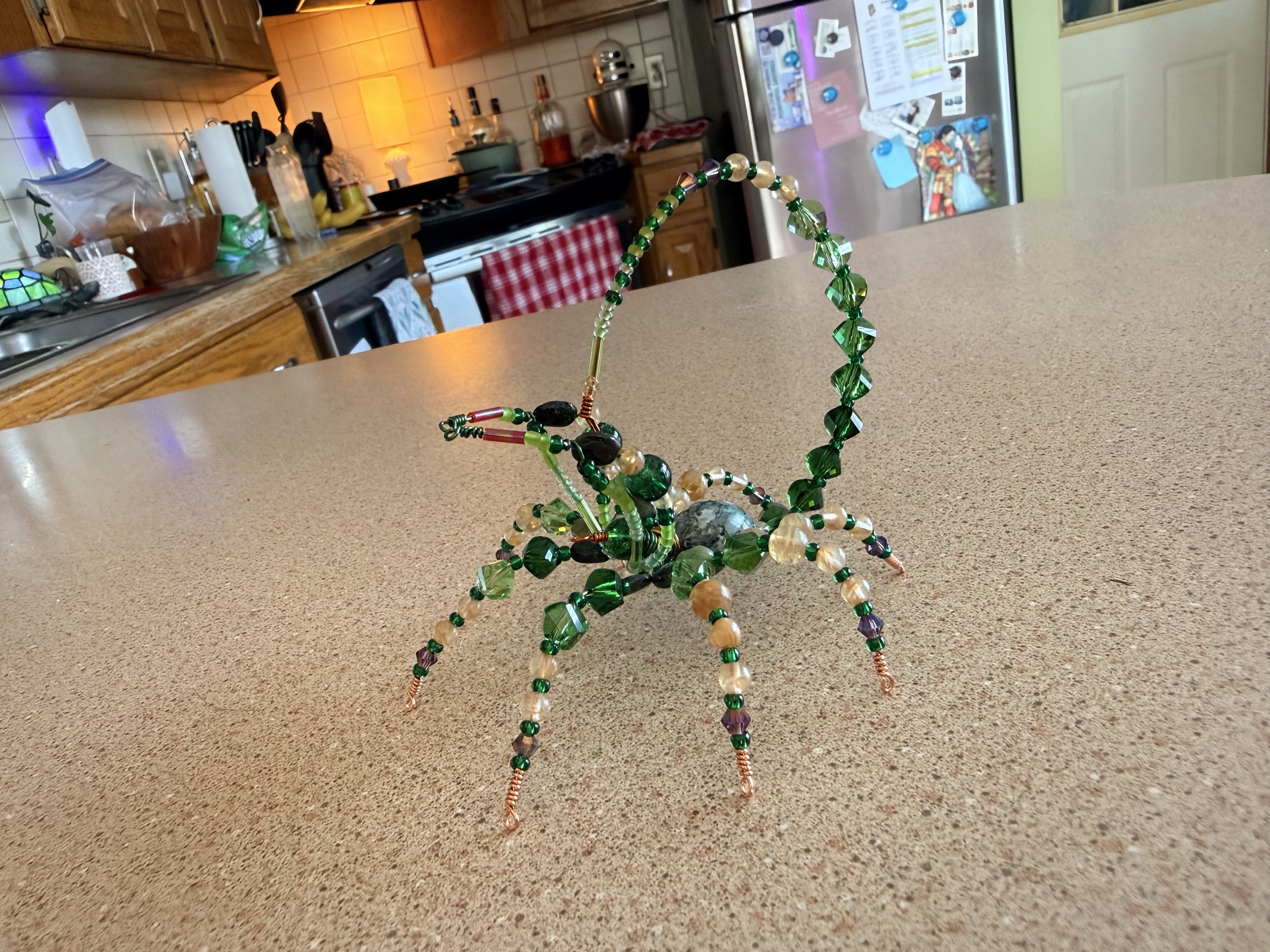 Colorful beaded scorpion on a beige countertop in a kitchen with wooden cabinets, a tiled backsplash, and a stainless steel refrigerator with various photos and notes attached.