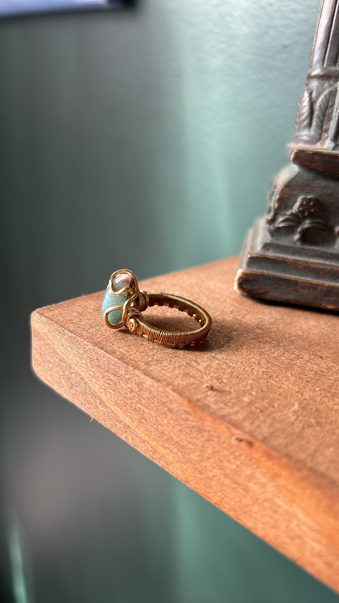 A gold wire ring with a blue stone sitting on a wooden surface. Part of a decorative object with carved details is partially visible on the right side.