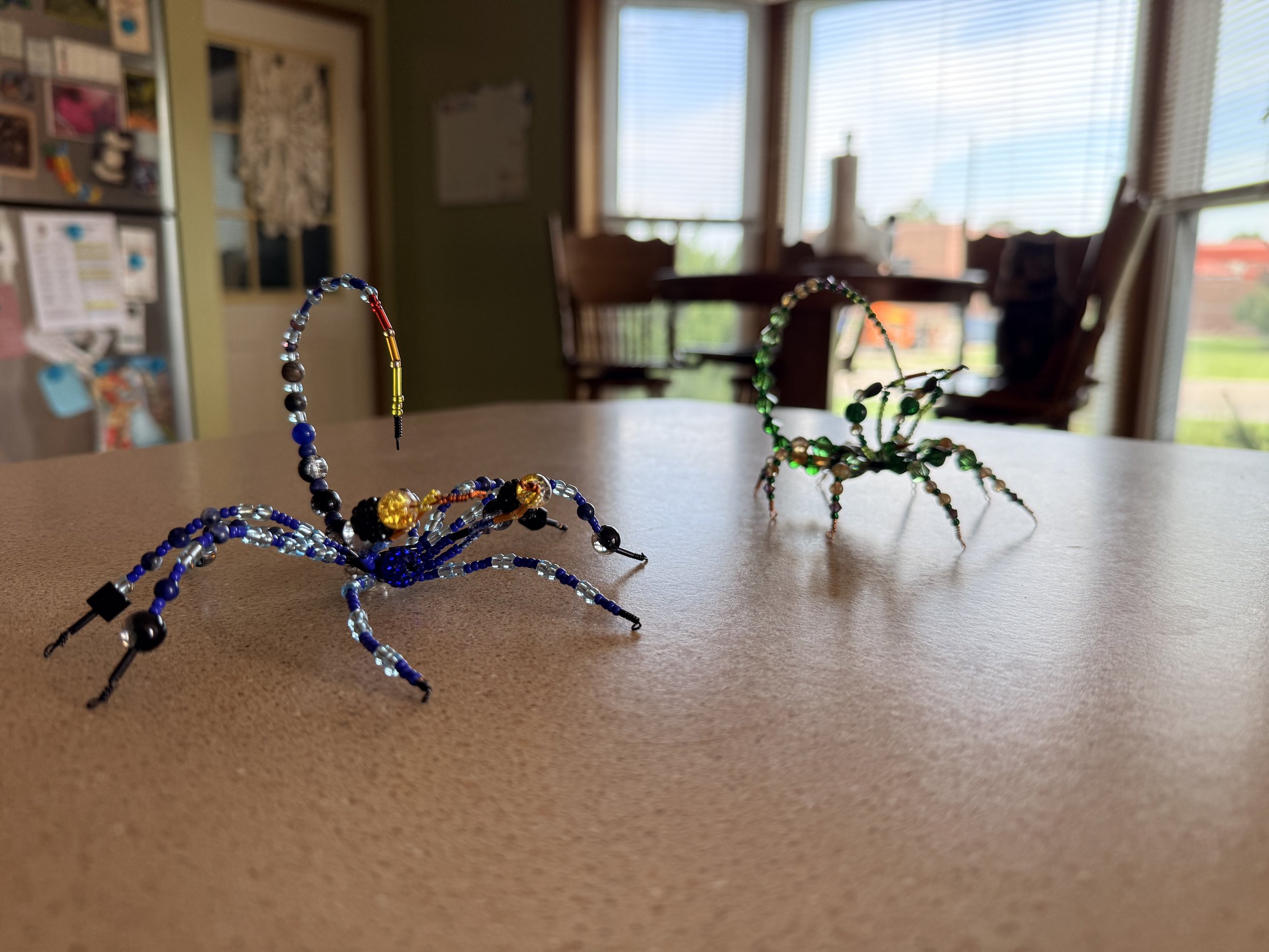 Colorful beaded scorpions  on a table in a bright, sunny room with large windows and wooden chairs.
