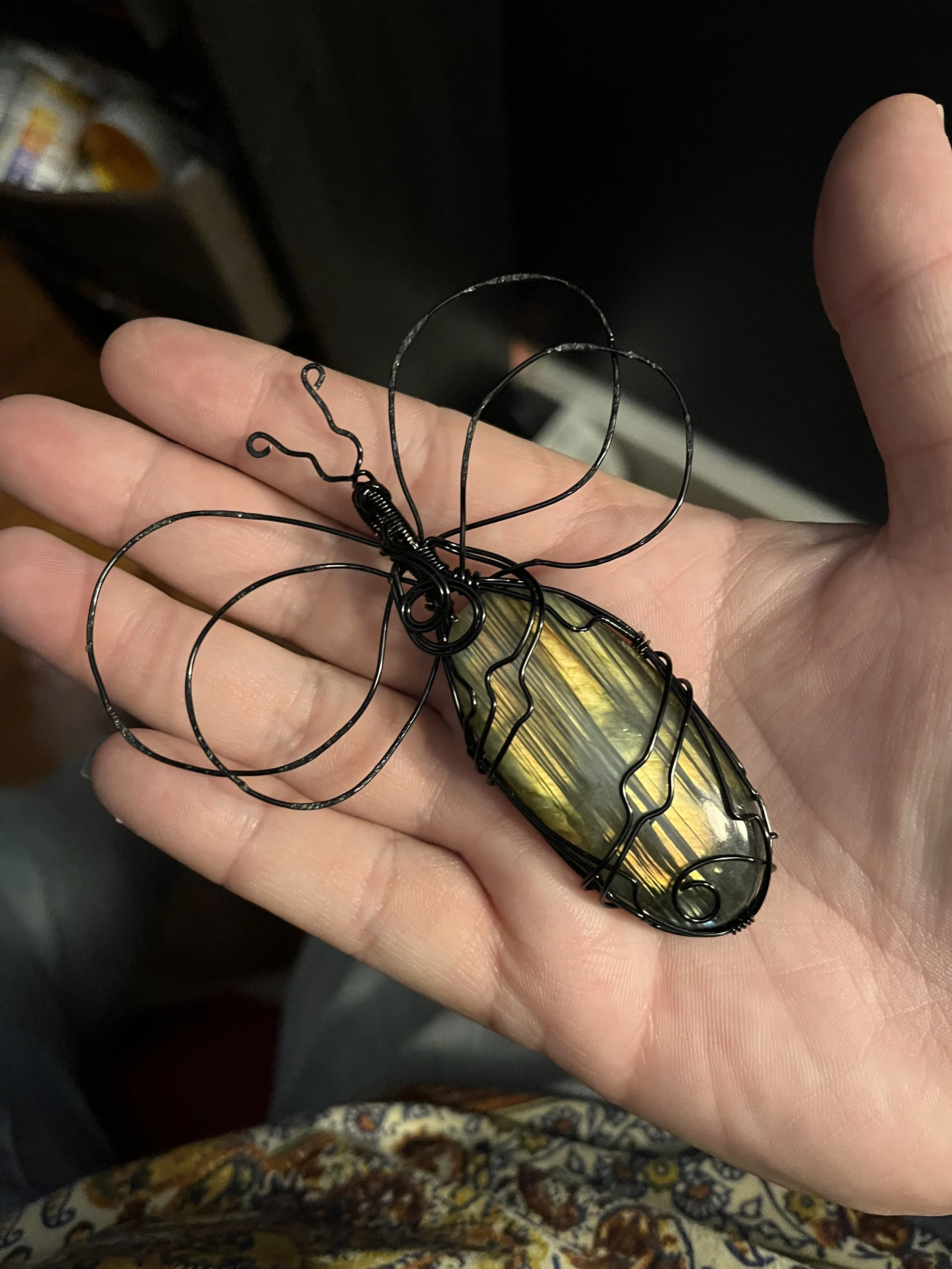 A person's hand holding a decorative wire butterfly with a yellow and black striped stone body. The background includes a dark setting with some blurred objects.