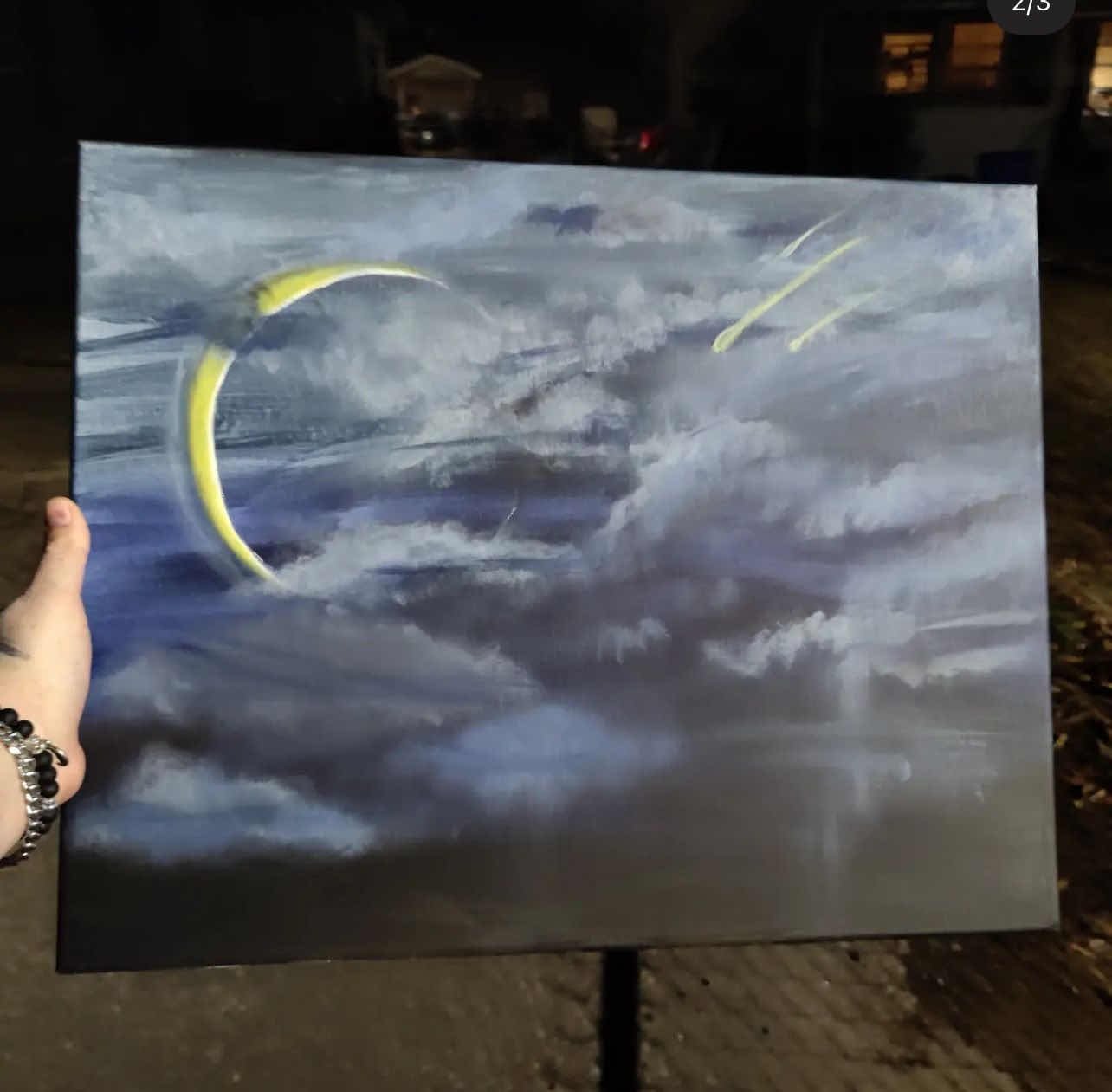 Nighttime outdoor scene of a hand holding a painting of a cloudy sky with a crescent moon and streaks of light.