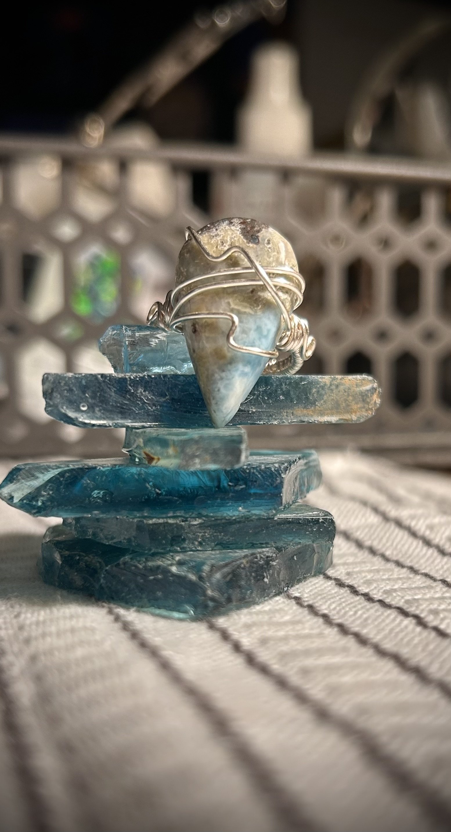 Close-up of a blue and beige gemstone ring wrapped with silver wire, placed on stacked irregular blue glass pieces with a blurred background of white bottles.