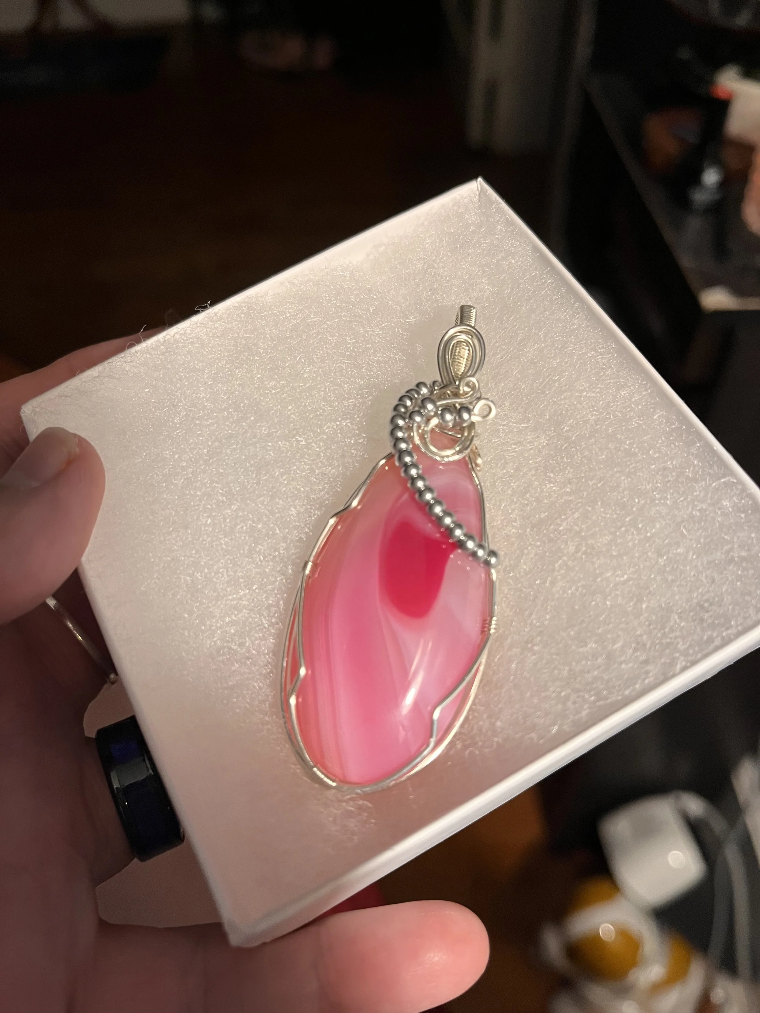 A person holding a white box with a pink and irregularly shaped gemstone pendant attached to a silver ball chain necklace. The background includes shelves with other jewelry items.