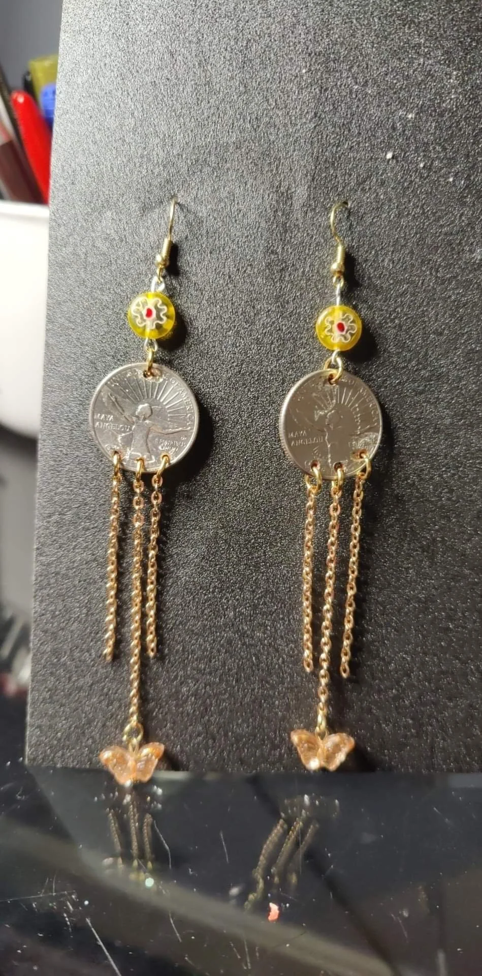 Pair of gold-colored earrings featuring a small yellow flower bead at the top, a USD quarter coin in the middle, with three gold chains hanging below, ending in a small butterfly-shaped charm.