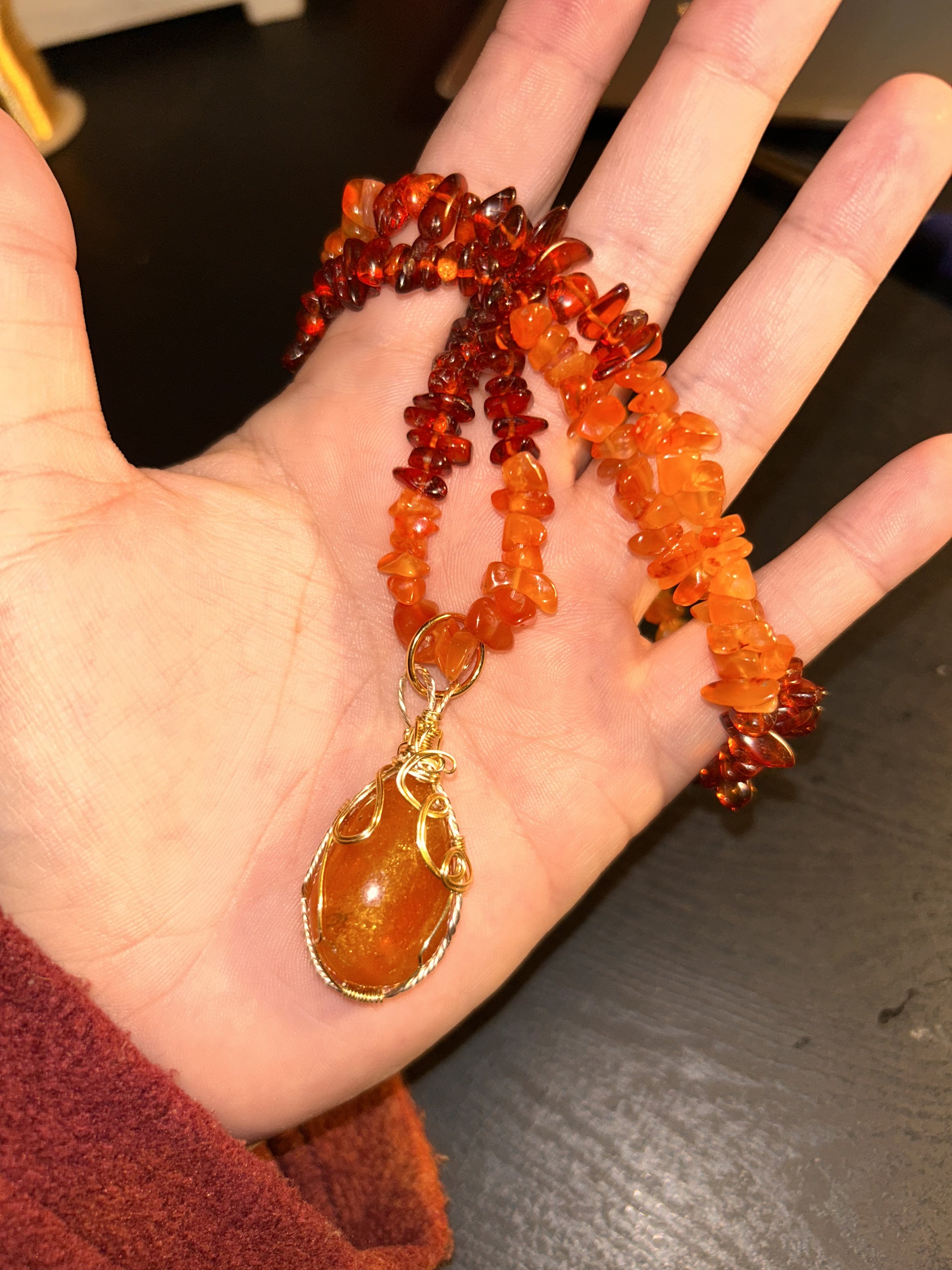 A person's hand holding two strands of orange and red gemstone beads with a large amber pendant attached.