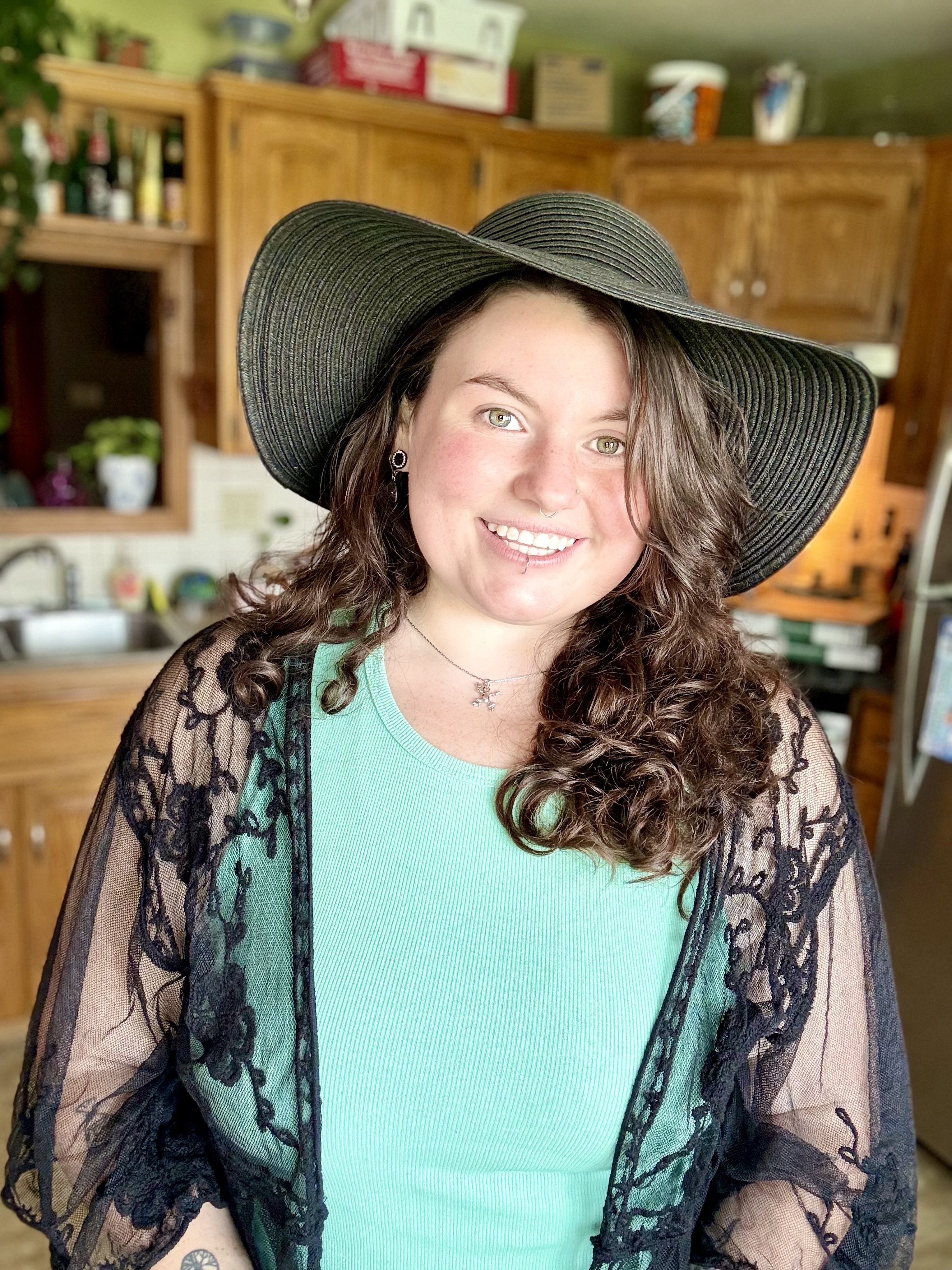 A young woman with curly brown hair and light skin smiling, wearing a large black sun hat, a turquoise top, and a black lace shawl, standing in a kitchen with wooden cabinets and various items on the countertops in the background.
