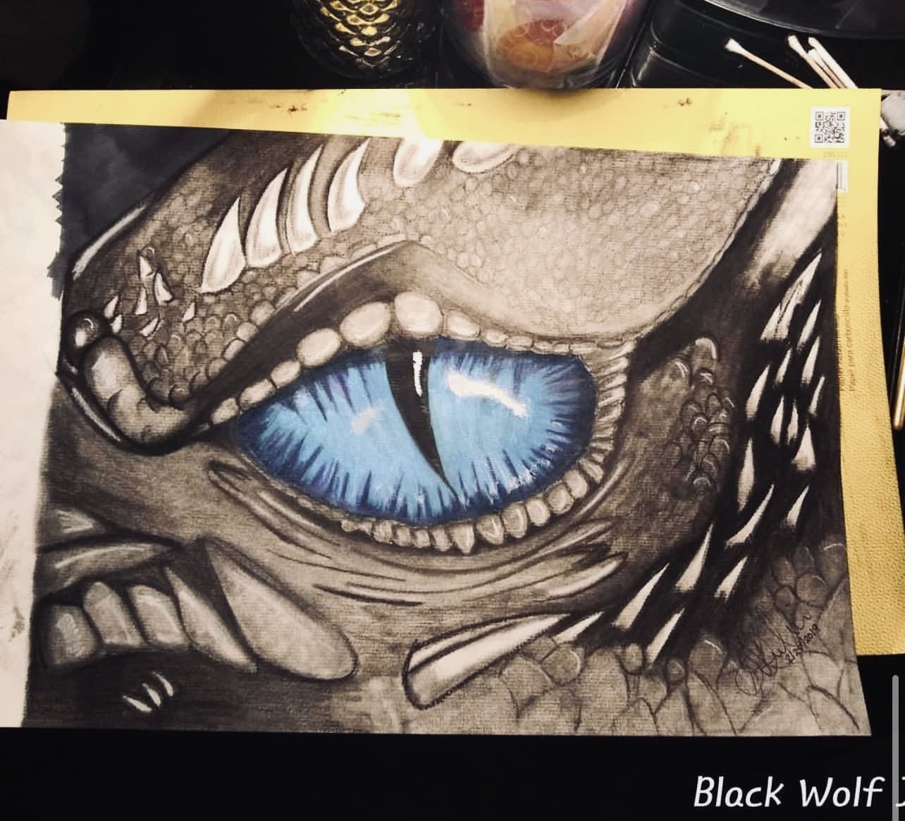 Close-up drawing of a dragon's face with a large blue eye and sharp teeth, rendered in black and white with detailed shading.