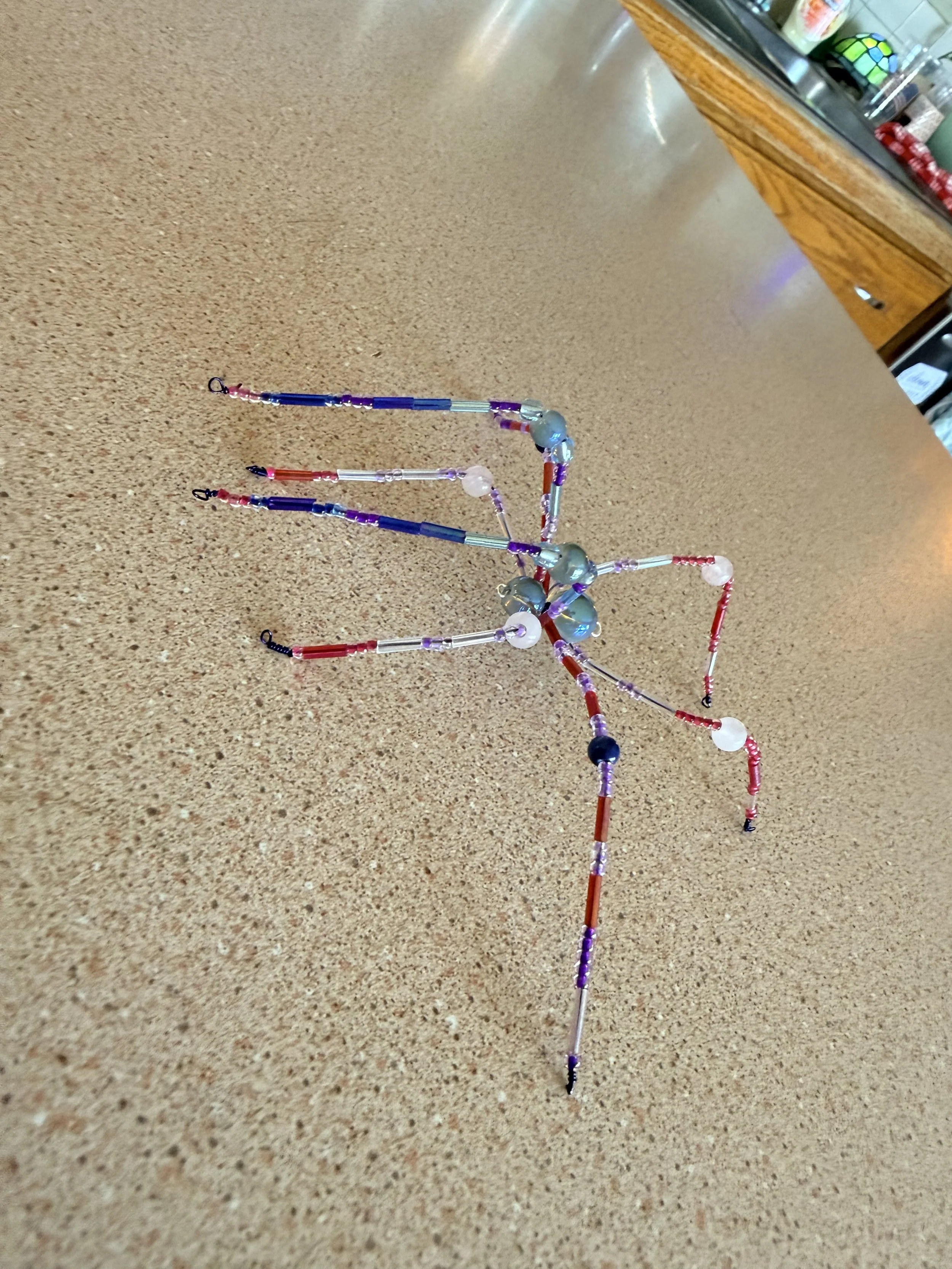 A colorful, bead and wire model resembling a spider or insect on a beige countertop, with kitchen items in the background.