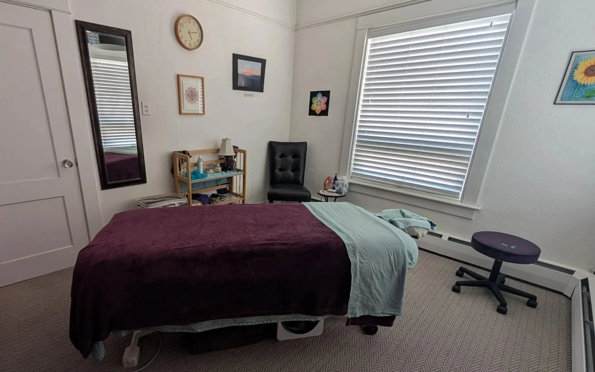 A safe and inviting massage treatment room in a spiritual, South Denver massage studio. White walls with natural light, hand-drawn art, and a cozy table with warm flannel sheets