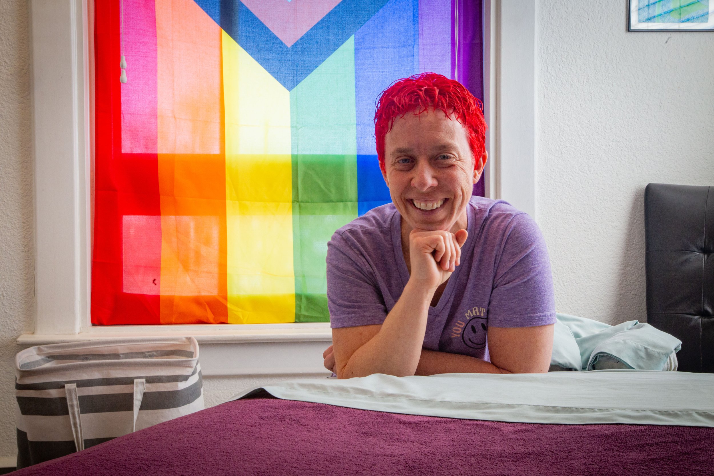 headshot of a smiling Julie, owner and massage therapist of Ember & Flow bodywork and massage studio. She is wearing a purple cotton tee shirt with vibrantly red hair,  leaning on her massage table, perched in front of a colorful pride flag
