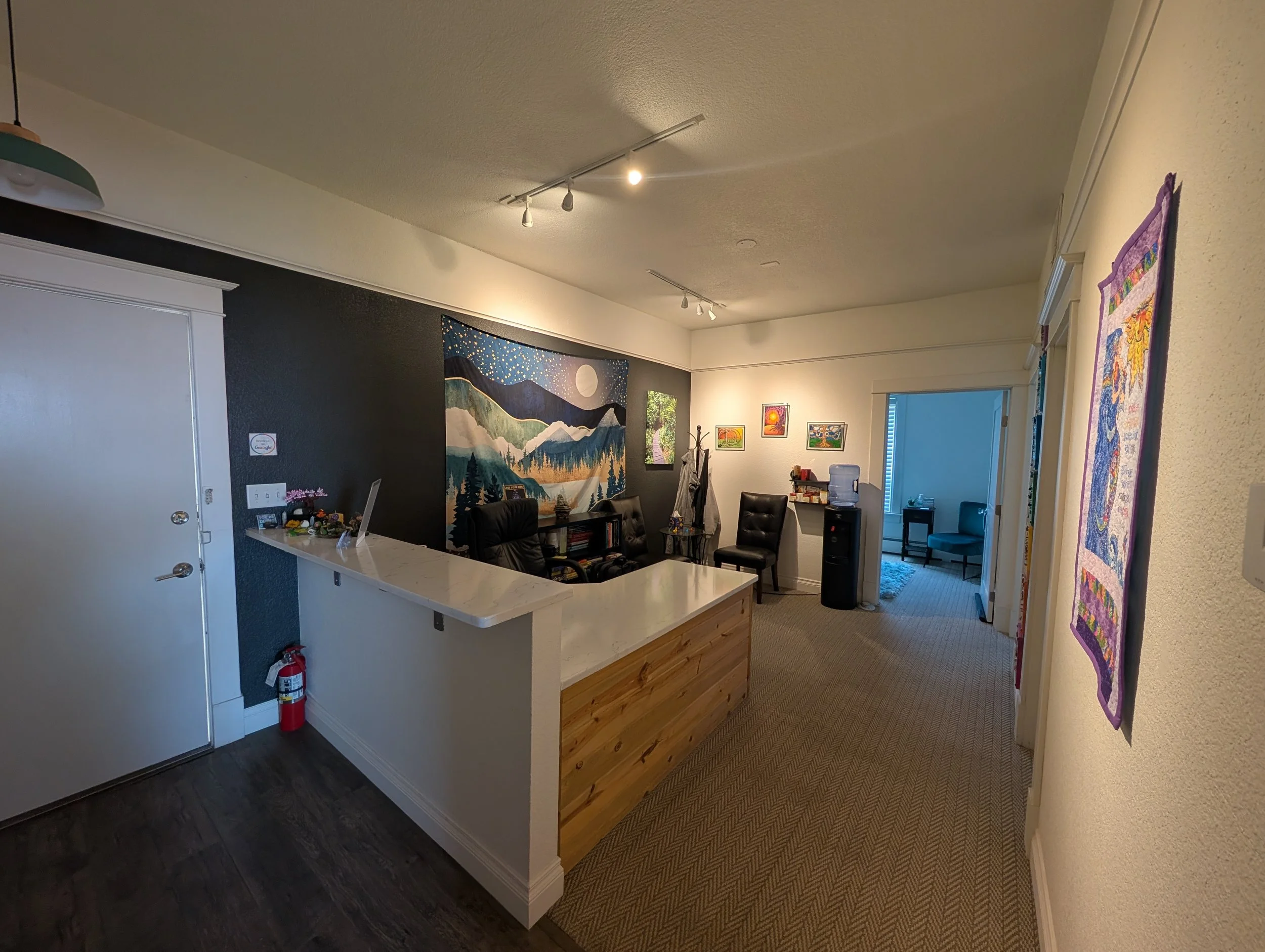An inviting waiting room with a reception desk, a mountain and night sky mural, and framed spiritual / nature artwork. There's a white reception desk, chairs and a water cooler.
