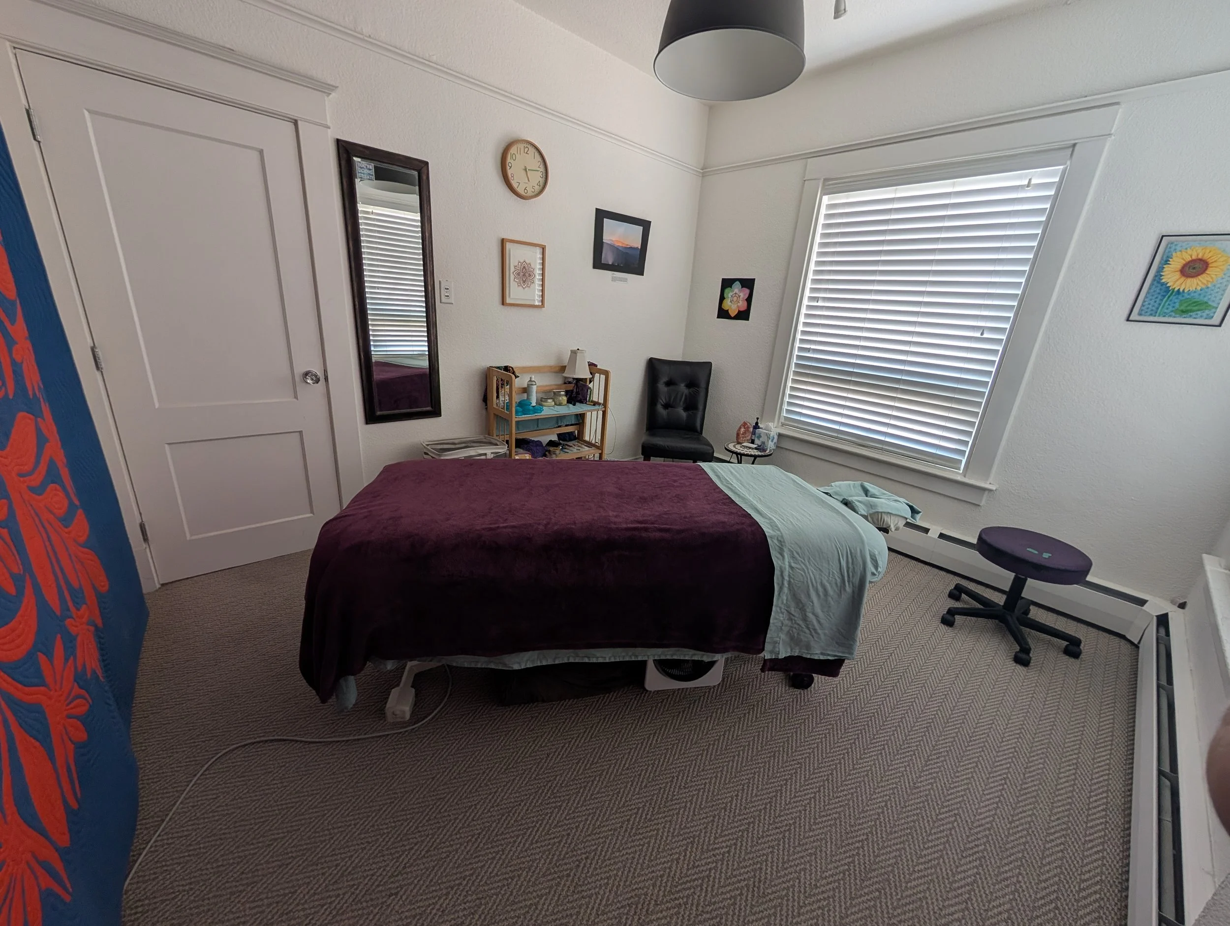 An inviting massage therapy room within this safe, South Denver bodywork studio. Photo features an inviting massage table covered in warm sheets and a purple comforter, with natural light, and Julie's own hand-drawn art.