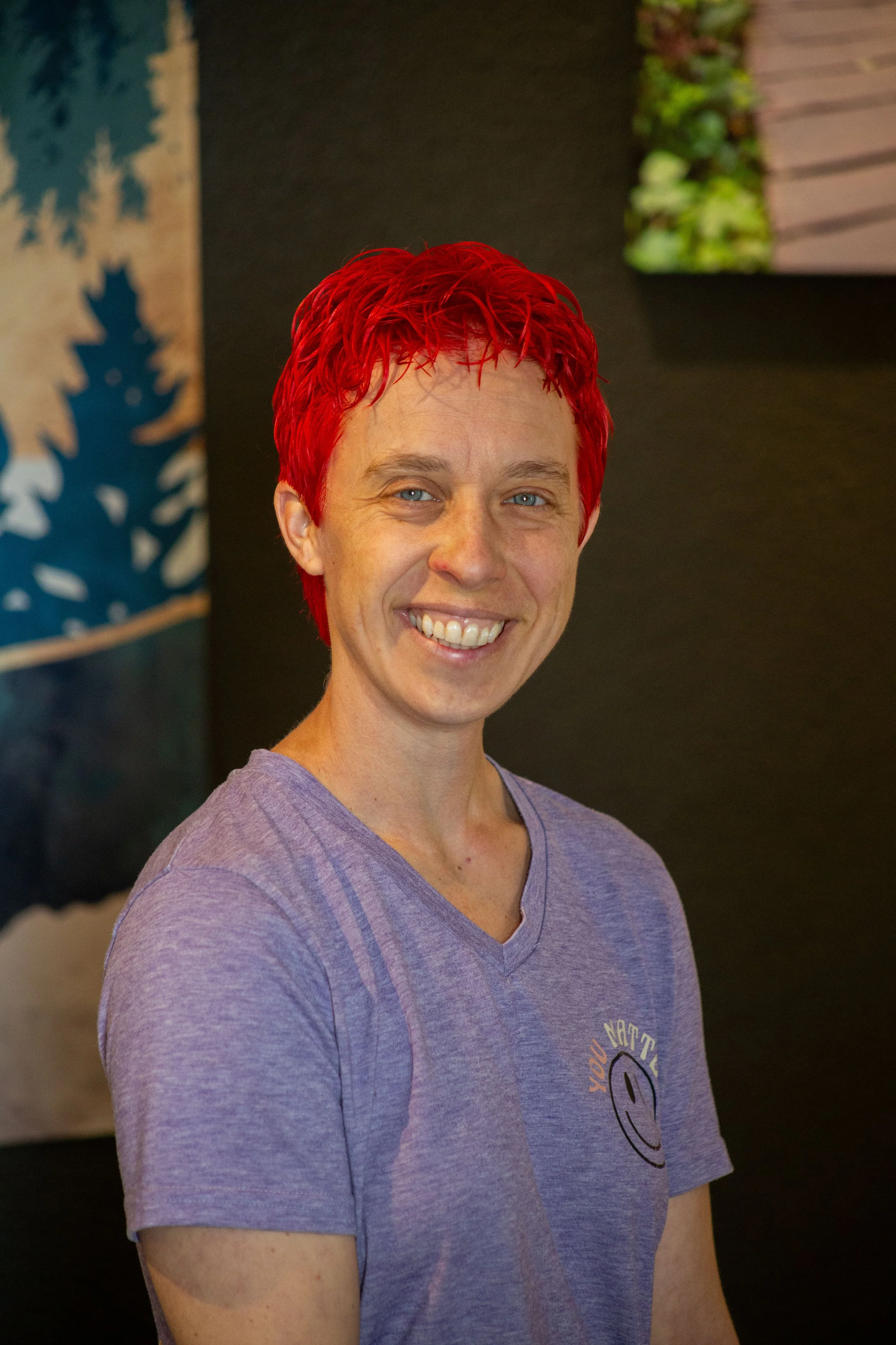 portrait shot of Julie, intuitive massage therapist, smiling at the camera with vibrantly red hair, wearing a purple cotton tee shirt, in her South Denver massage studio