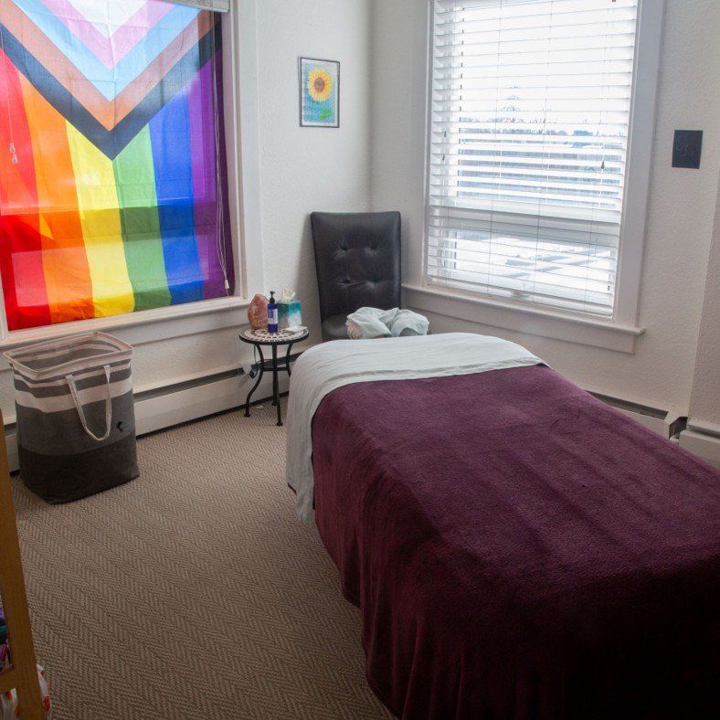 The Ember & Flow massage studio in South Denver operates out of a clean, inviting massage therapy room with natural lighting, a pride flag, and a comfortable massage therapy table draped in cozy sheets