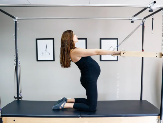 Pregnant woman performing Pilates exercise on a reformer machine in a studio.  Logo for a wellness friend that this massage therapy studio endorses.