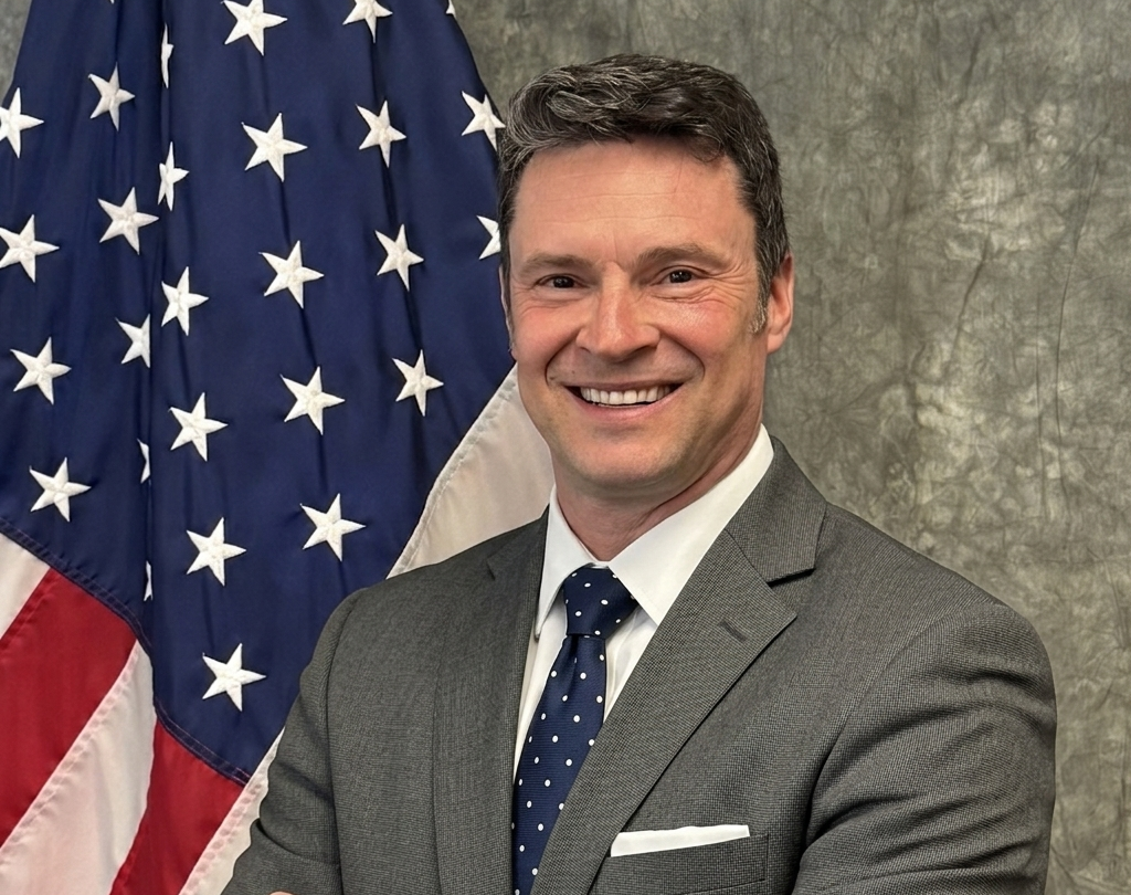 A man in a gray suit, white shirt, and navy polka dot tie standing in front of an American flag, smiling.