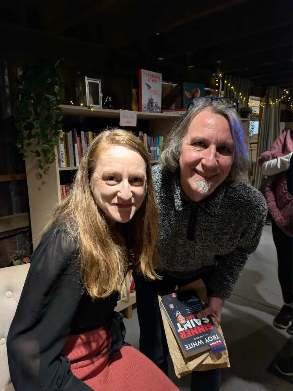 Two people, a woman and a man, at a bookstore or library. The woman has long red hair and is wearing a black top with a red skirt. The man has shoulder-length gray hair, glasses resting on his head, and a gray fuzzy sweater. They are smiling and the 