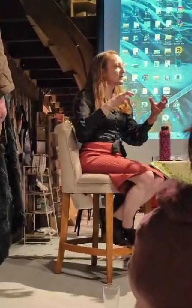 A woman with red hair and a black shirt is sitting on a stool, gesturing with her hands, possibly giving a presentation or speech. Behind her is a large screen with icons from a computer desktop. The setting appears to be indoors, with wooden stairs 