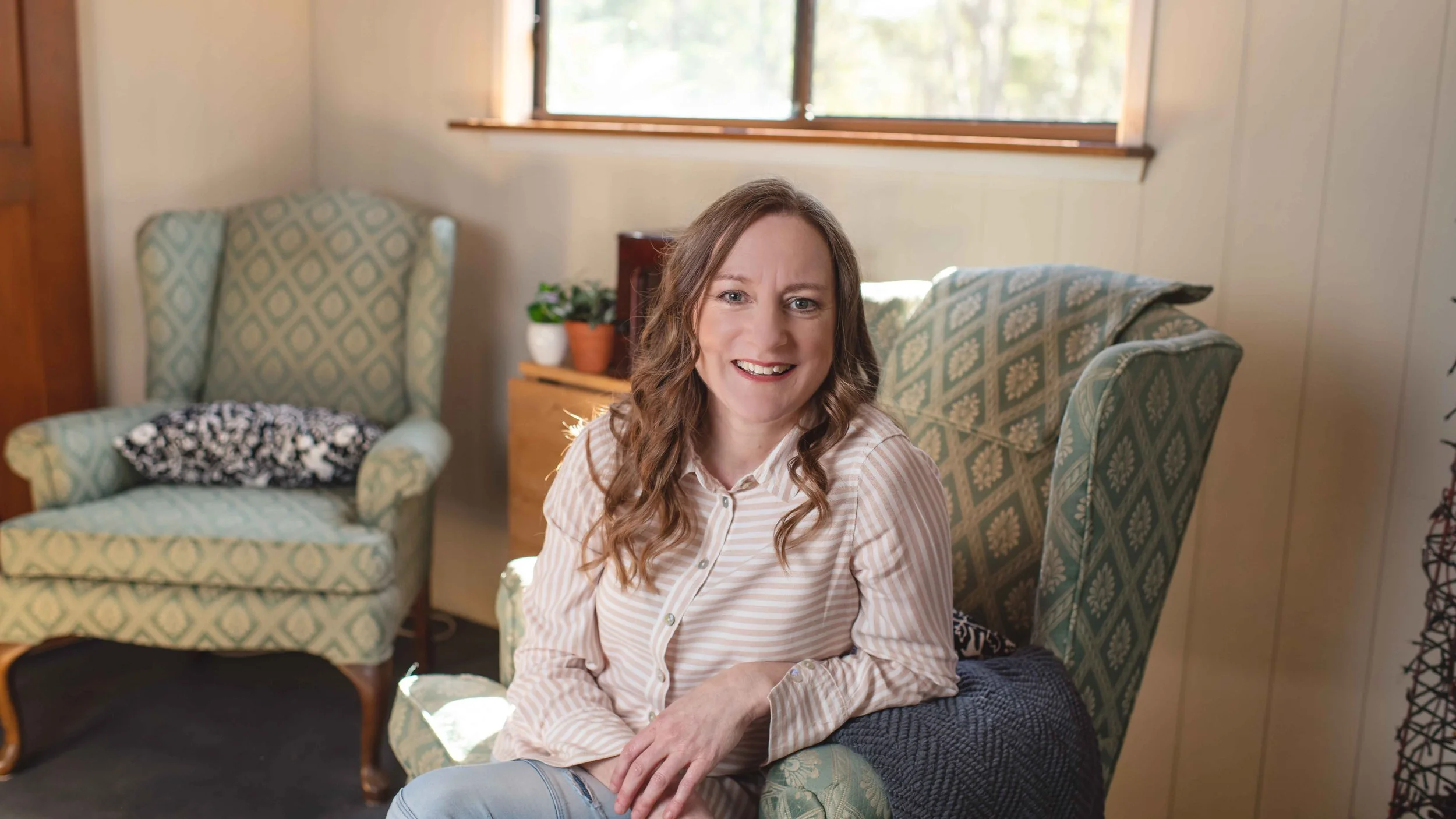 Author Clare Bruce smiling while sitting on a green patterned armchair in a cozy living room with sunlight coming through a window behind her.