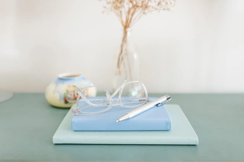 A pair of clear eyeglasses and a silver pen resting on two stacked books, on a light green table with a decorative vase and dried flowers in the background.