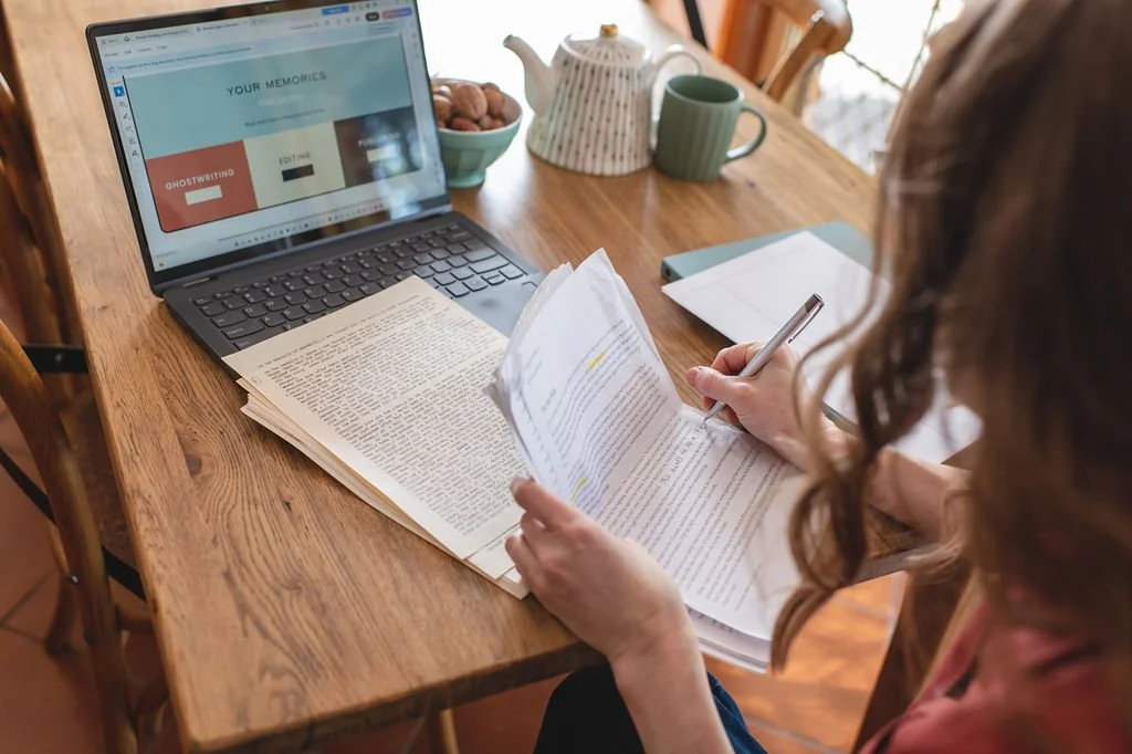 Author Clare Bruce sitting at a wooden table with an open book and writing on a piece of paper. There is a laptop, a teapot, a mug, and notebooks on the table.