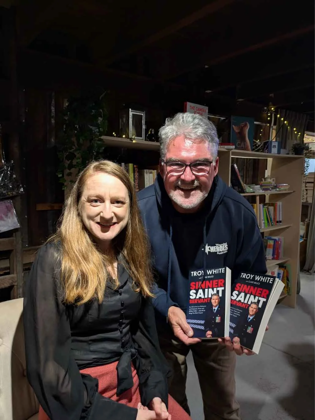 A woman and a man smiling at a book signing event, with the man holding books titled 'Sinner Saint Servant'.