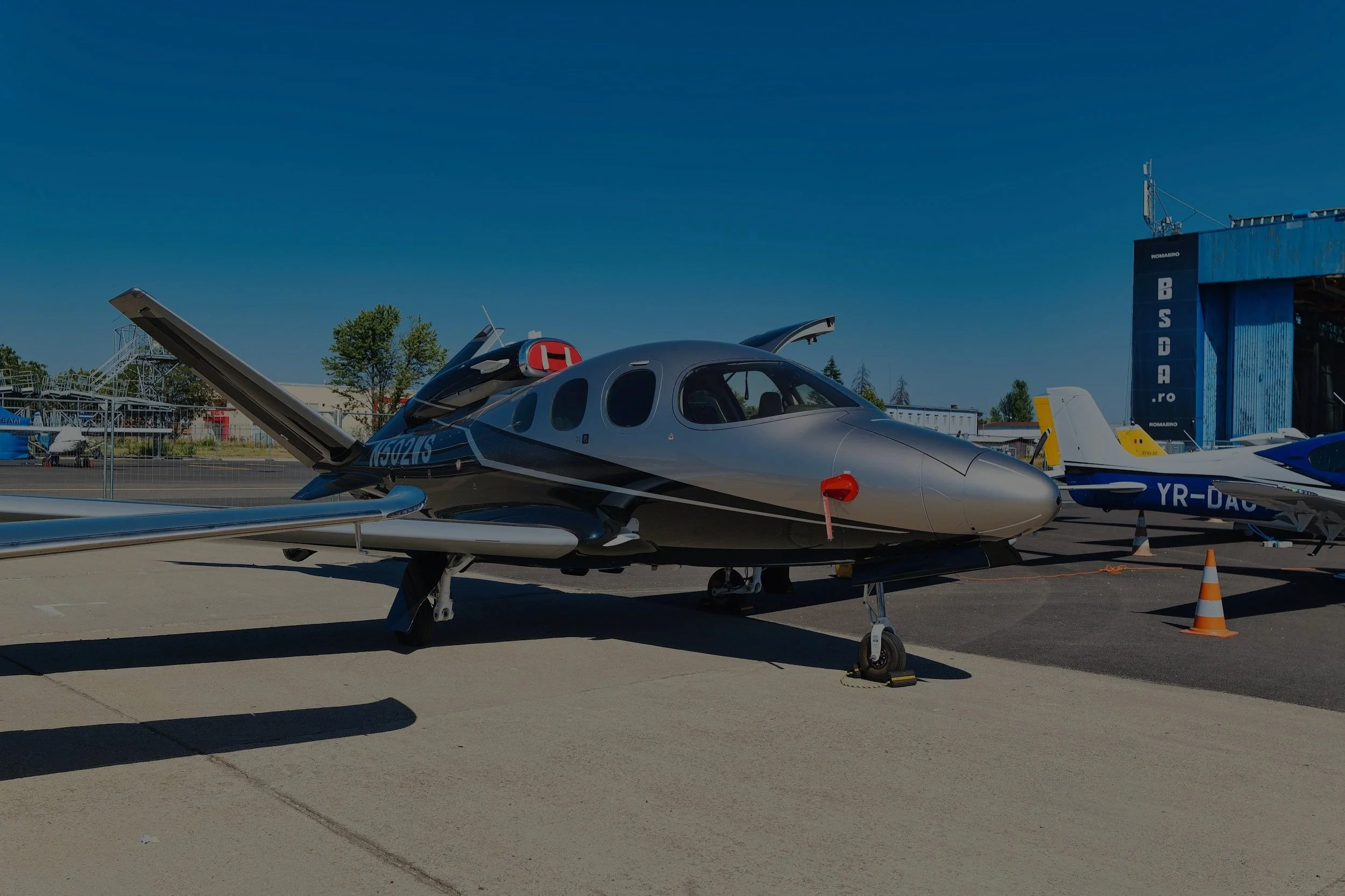 A small aircraft parked on an airfield with other vehicles and structures in the background, clear blue sky overhead.