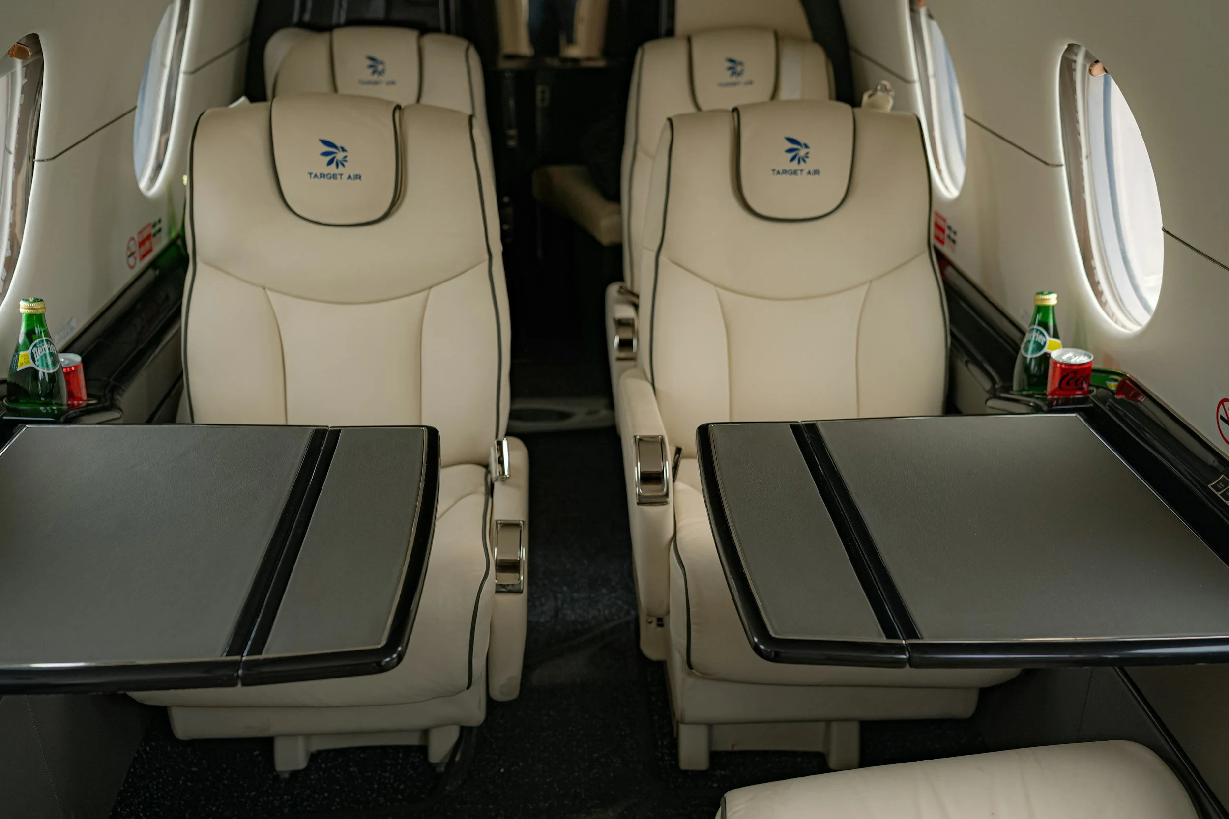 Interior of a Target Air airplane with cream-colored leather seats and foldable tray tables, with water bottles and soda cans on the sides.