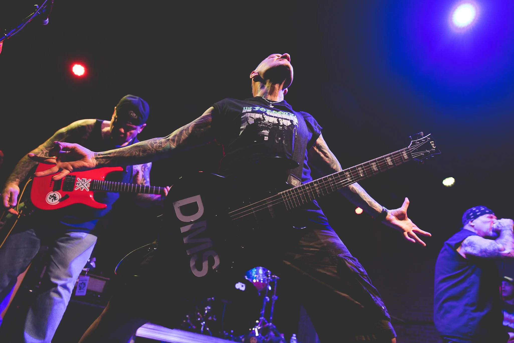 Musicians performing on stage with guitars, colorful stage lighting, and audience members visible.