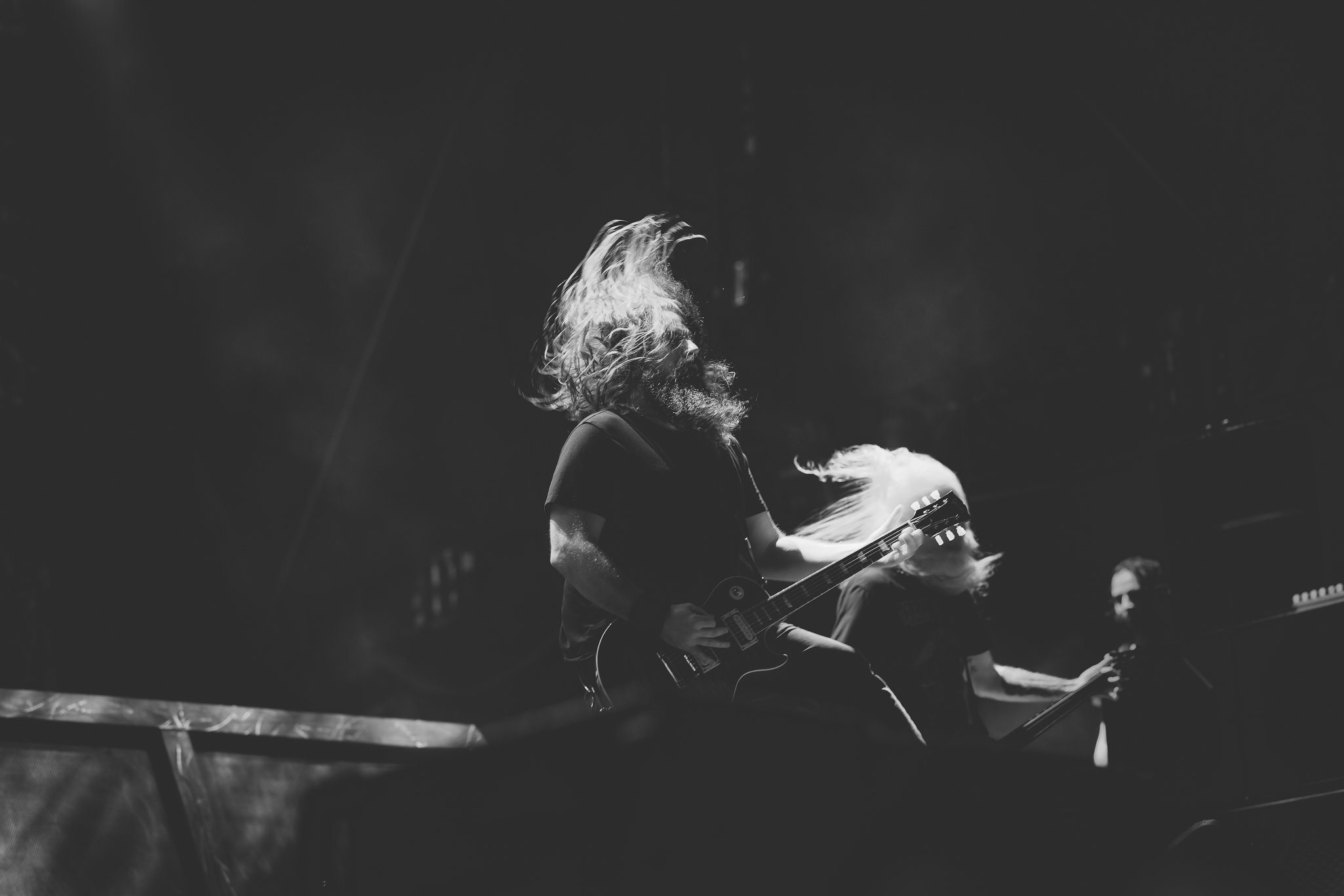 A black and white photo of a musician playing an electric guitar in a dark concert setting, with long hair and beard, focused on the instrument.