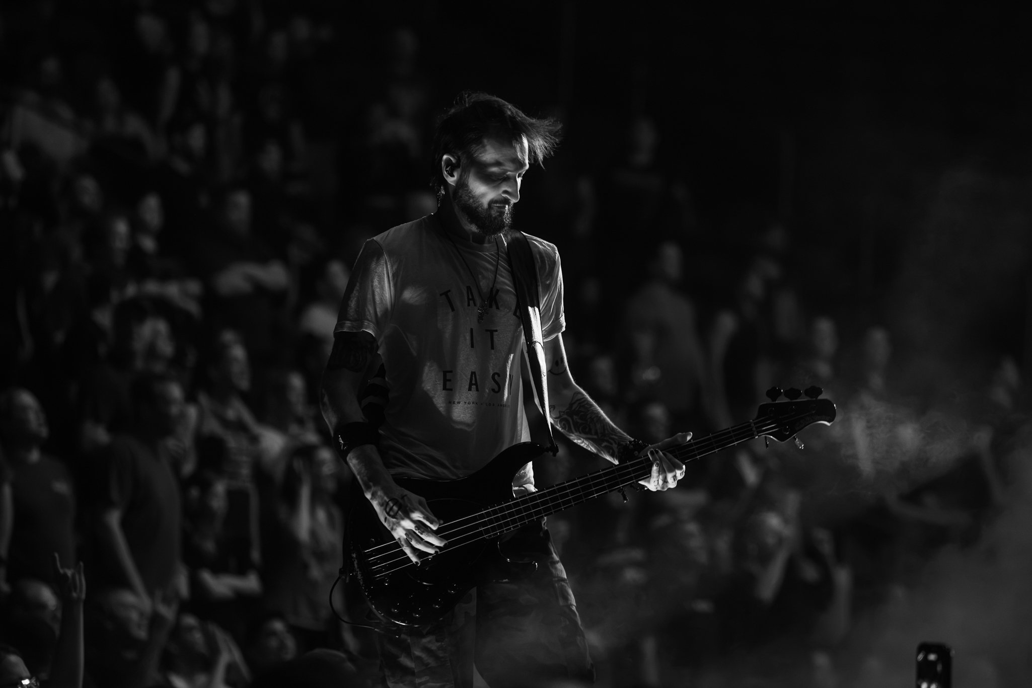 A musician with a beard and tattoos playing an electric guitar on stage, illuminated by a spotlight while surrounded by a blurred audience in the background.