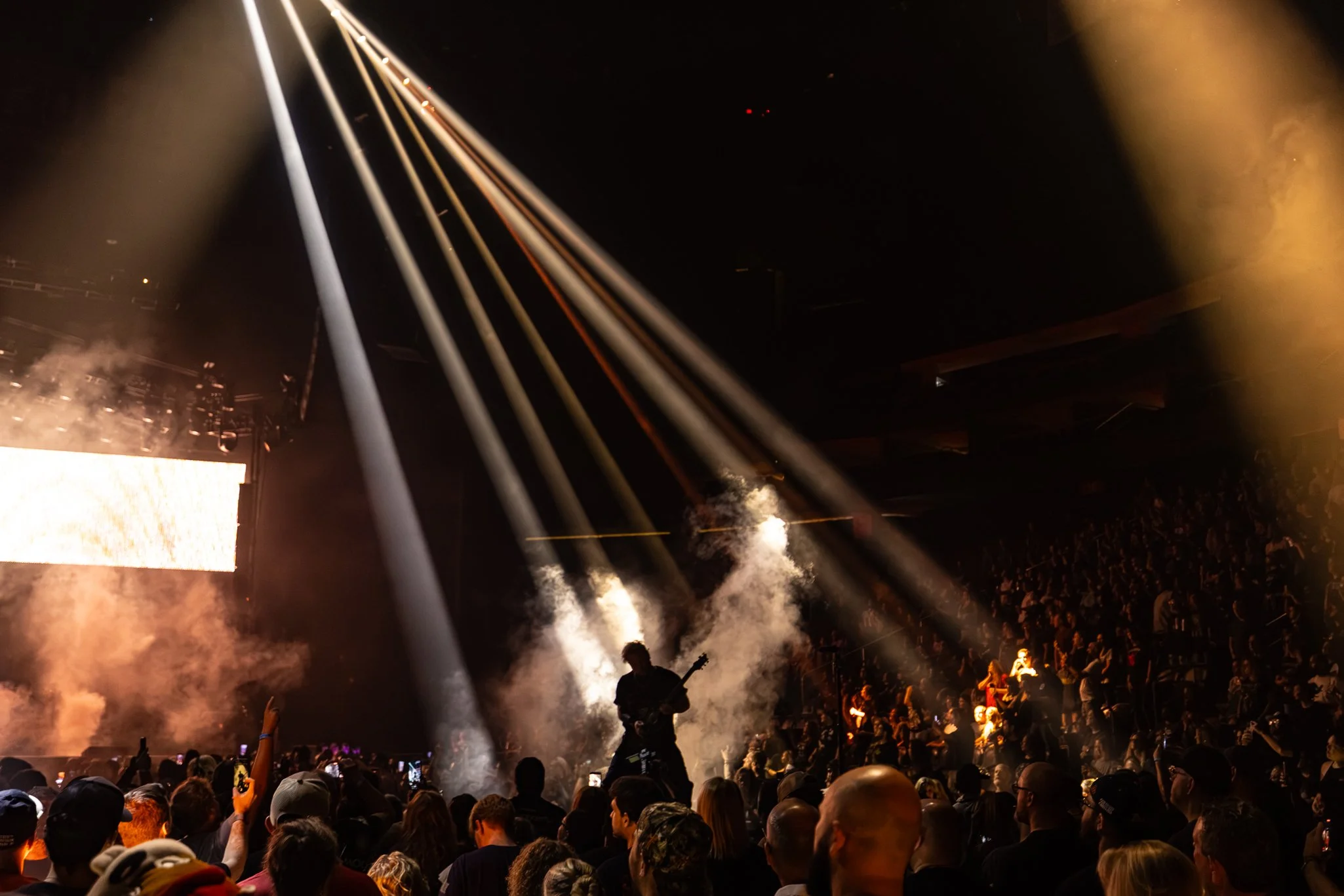 A concert or large event with an audience, stage, and bright beams of light shining into the dark sky. Smoke or fog is present, adding to the atmospheric effect, and people are taking photos or videos with their phones.
