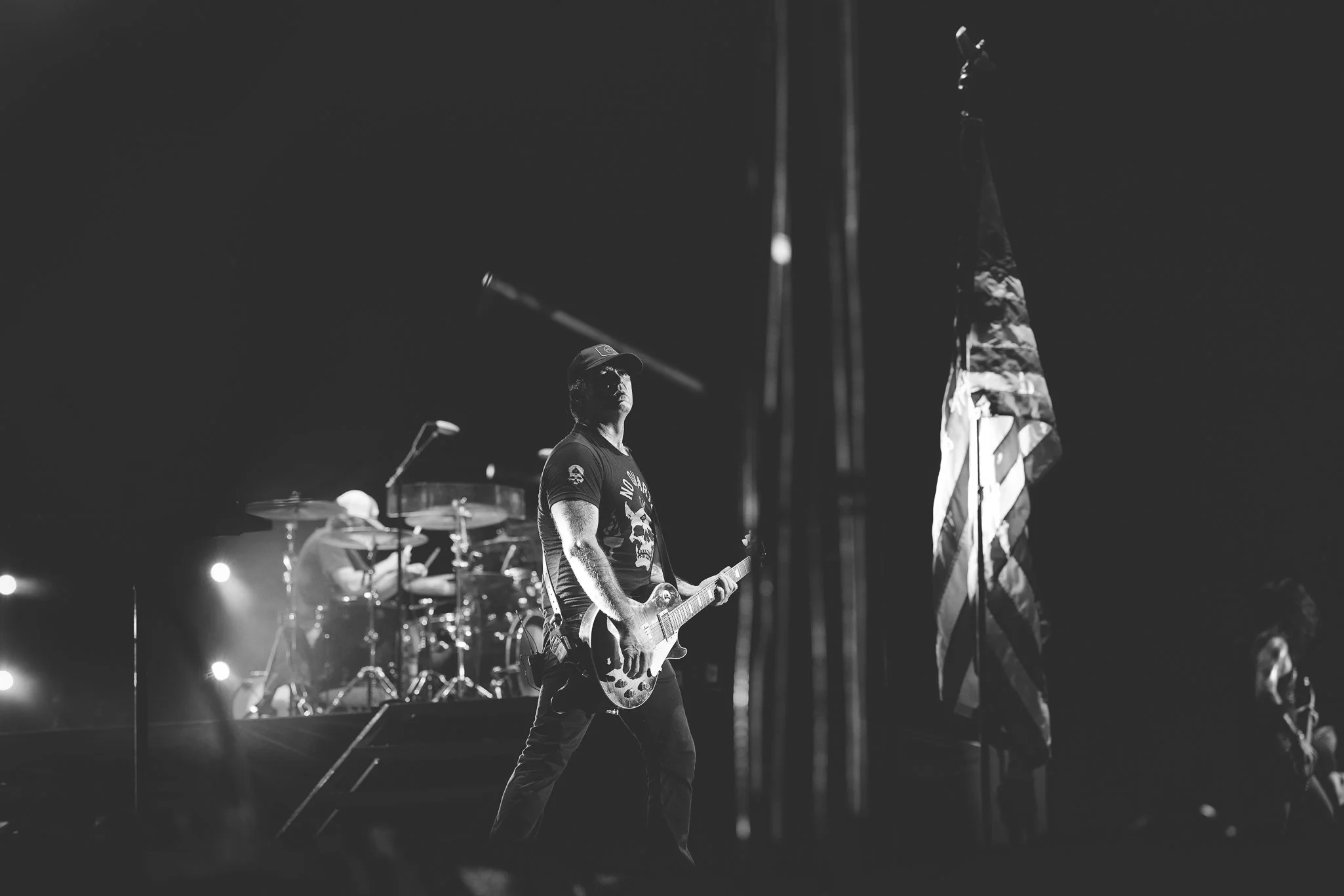 A black and white image of a musician playing an electric guitar on stage, with drums in the background and an American flag standing upright nearby.